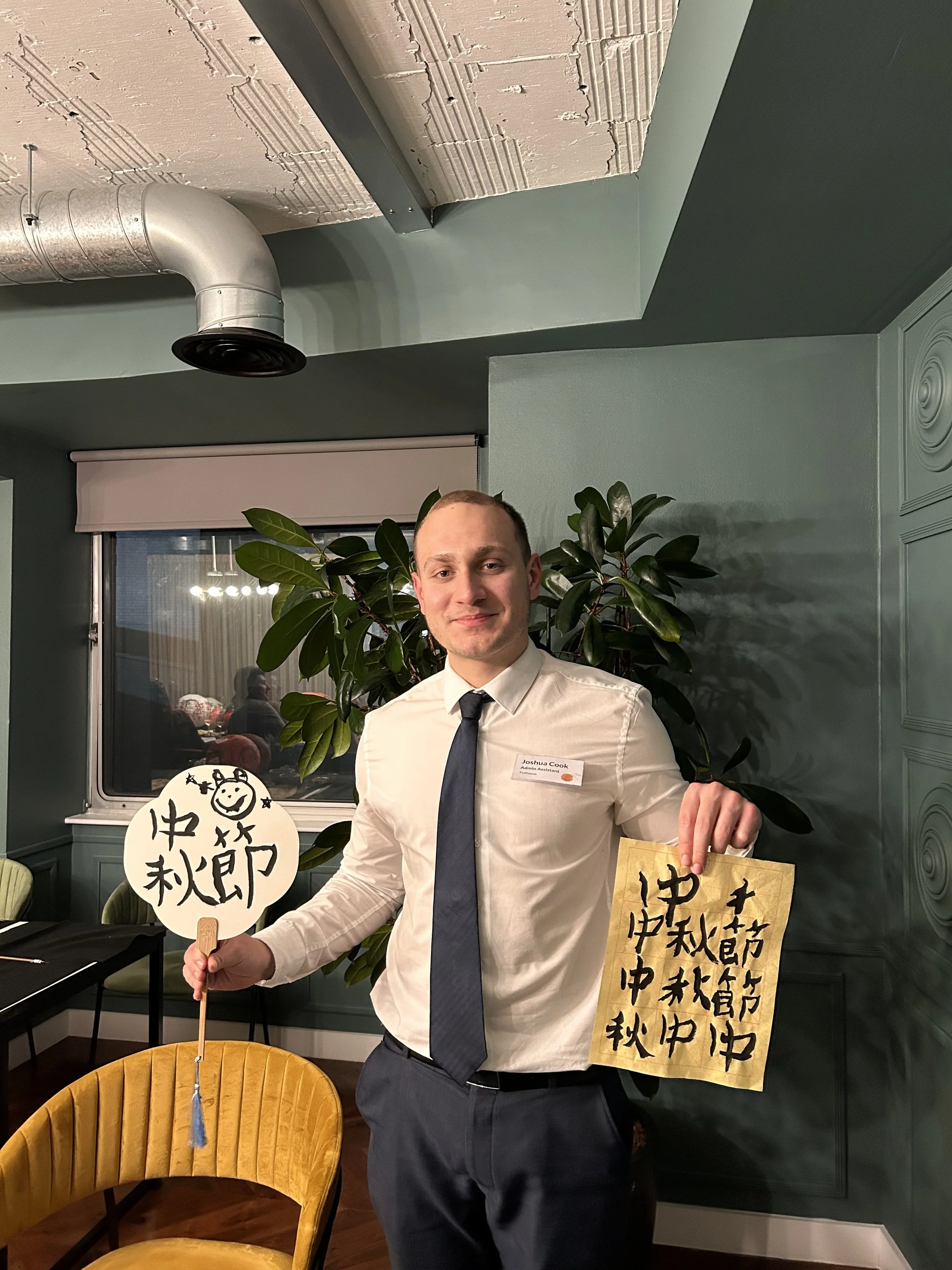 Company Workshop: Man in a tie holding a fan with Chinese characters and a paper with calligraphy, standing in a room with green walls and plants. workshop teacher: Yi Ling Lai, the founder of lingsspace