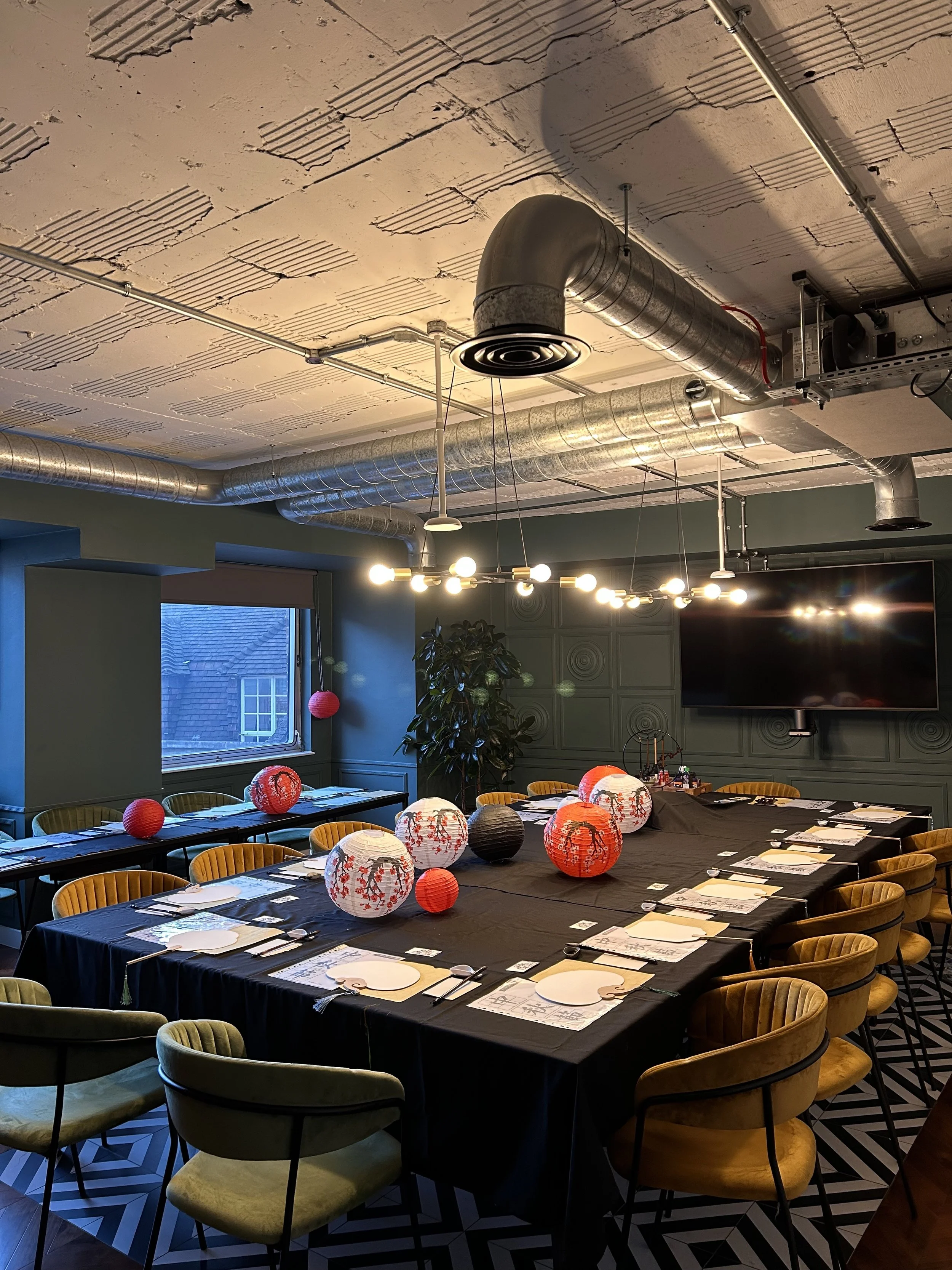 Company workshop display: A modern conference room with a large table set for a meal, decorated with colorful paper lanterns. The room features industrial-style ceiling with exposed ductwork, a geometric-patterned floor, and green cushioned chairs.