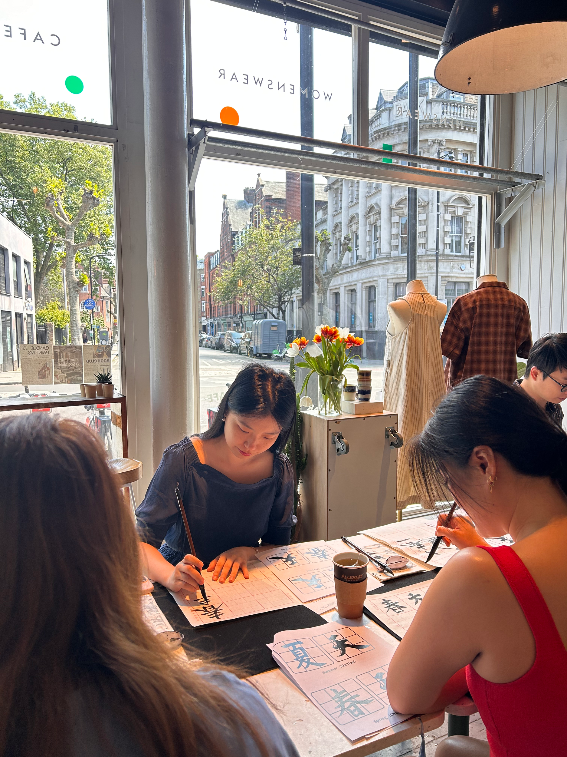 People seated at a long table practicing Chinese calligraphy on rice paper with brushes. Workshop teacher: Yi Ling Lai, the founder of lingsspace, Location: Aida, London