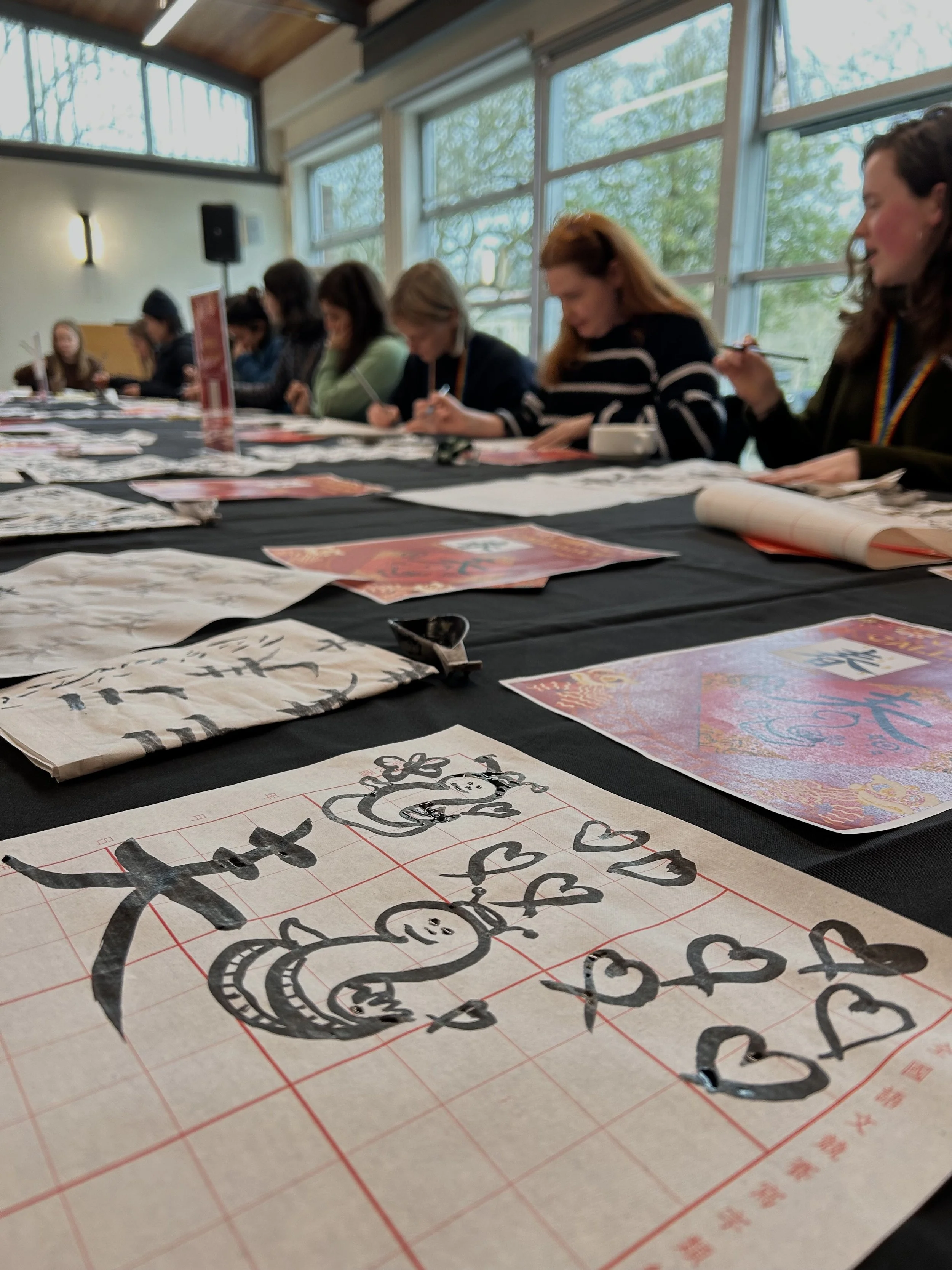 People participating in a calligraphy workshop, seated around a table with calligraphy paper and ink brush drawings. Workshop teacher: Yi Ling Lai, the founder of lingsspace, Location: Kew Garden London
