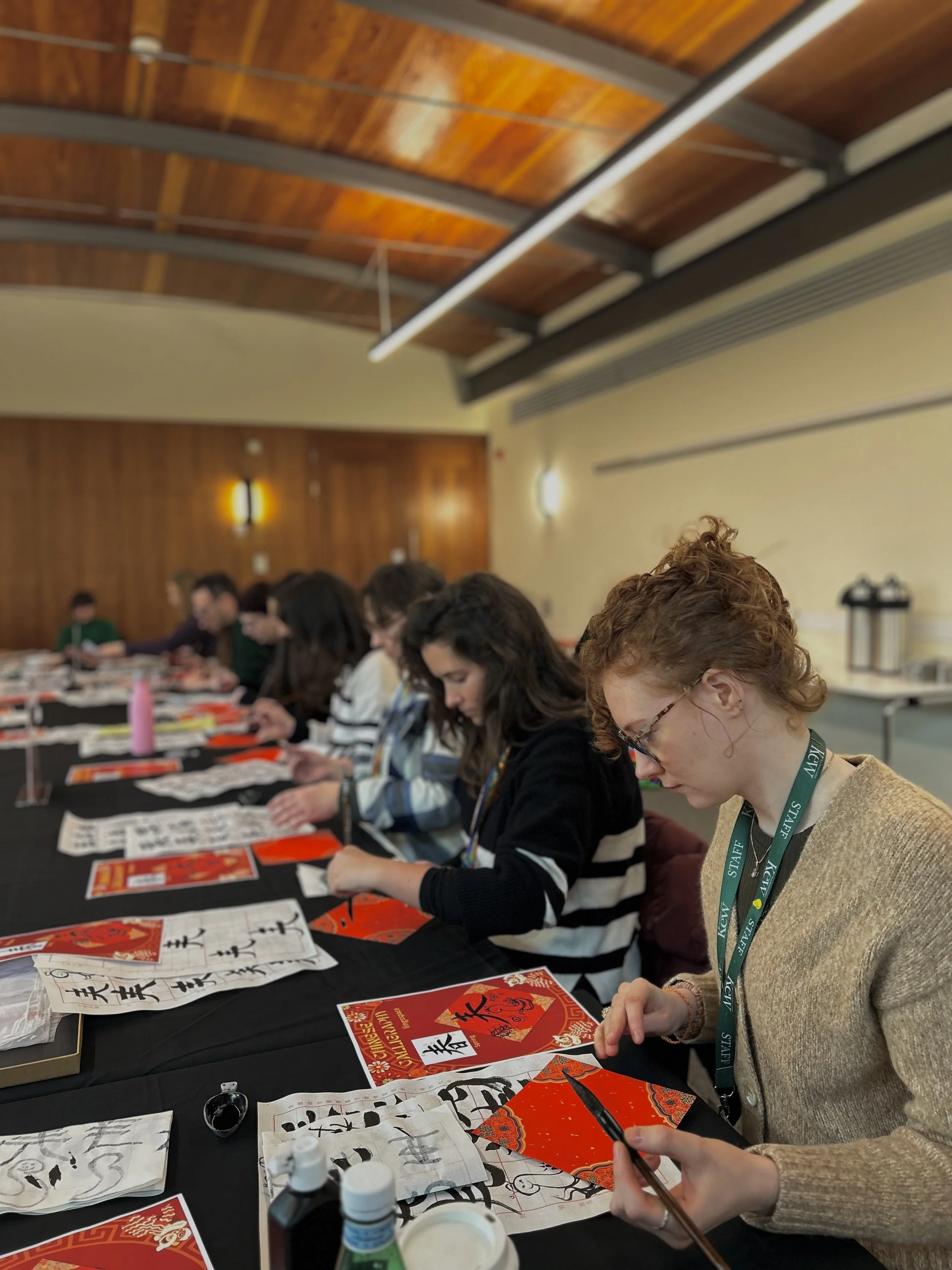 People seated at a long table practicing Chinese calligraphy on red paper with brushes, in a room with a wooden ceiling. Workshop teacher: Yi Ling Lai, the founder of lingsspace, Location: Kew Garden, London