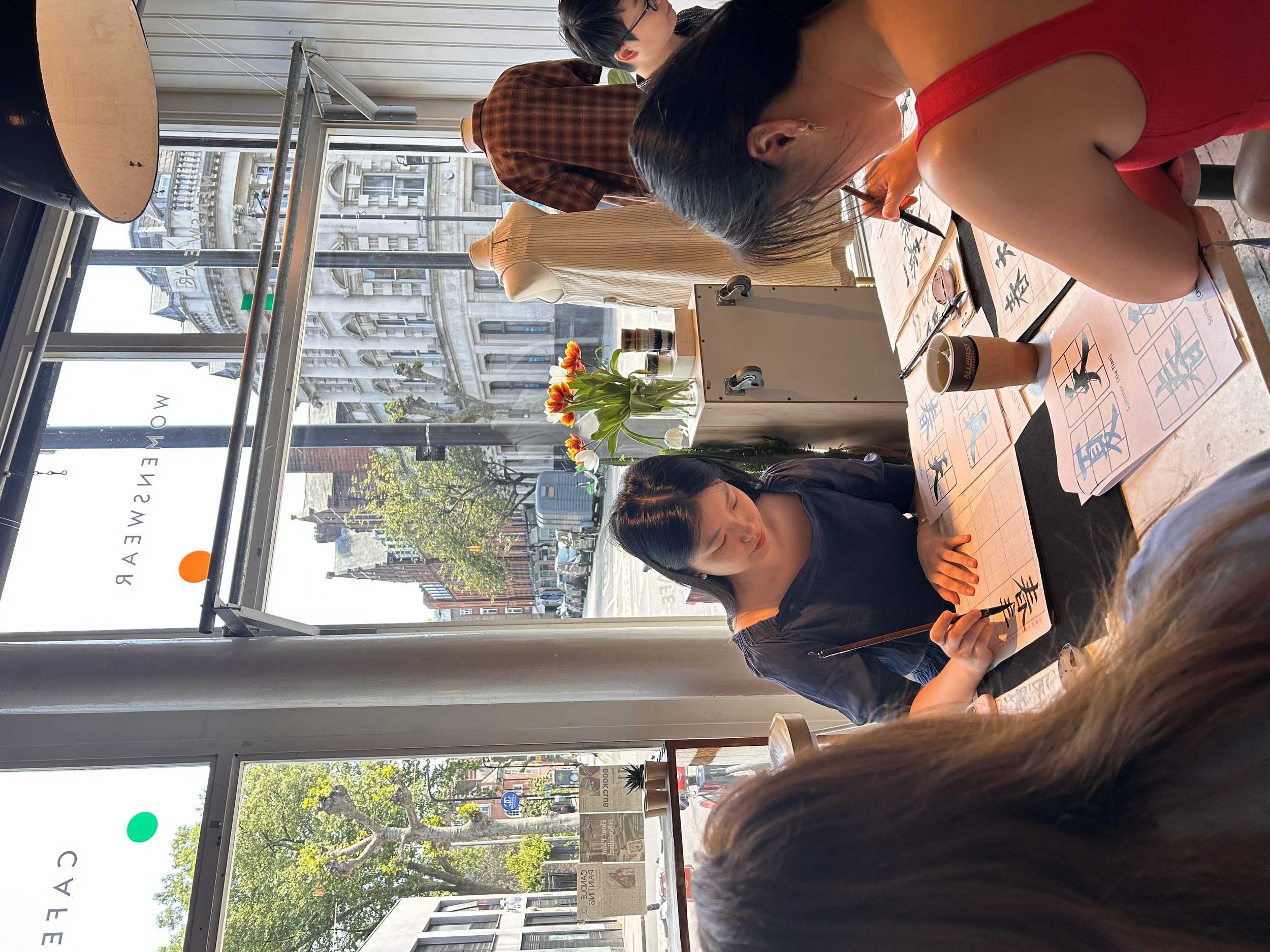 People seated at a long table practicing Chinese calligraphy on rice paper with brushes. Workshop teacher: Yi Ling Lai, the founder of lingsspace, Location: Aida, London