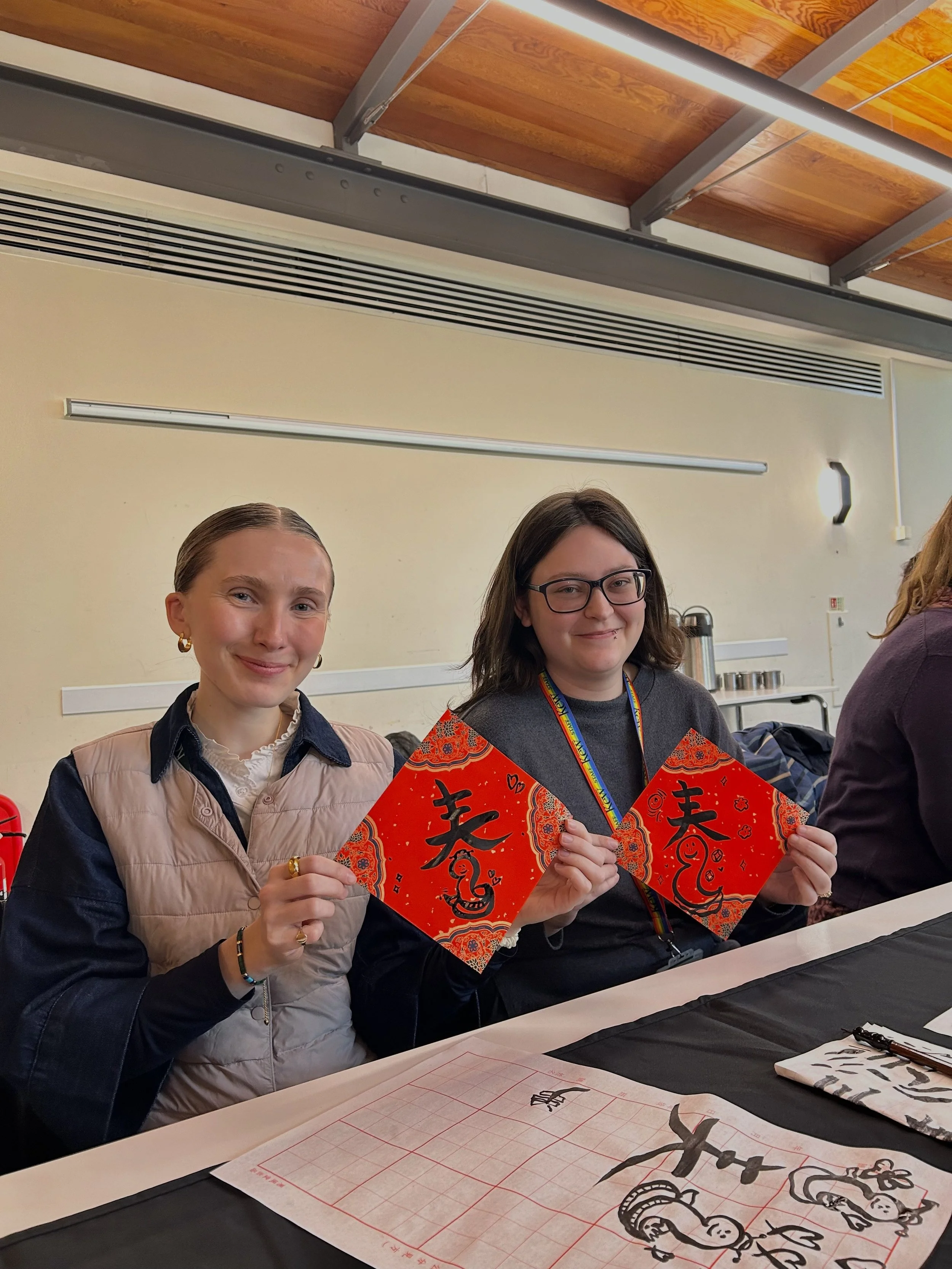 Two individuals sitting at a table, holding red paper squares with Chinese calligraphy. The table has calligraphy tools and paper. Workshop teacher: Yi Ling Lai, the founder of lingsspace, Location: Kew Garden London