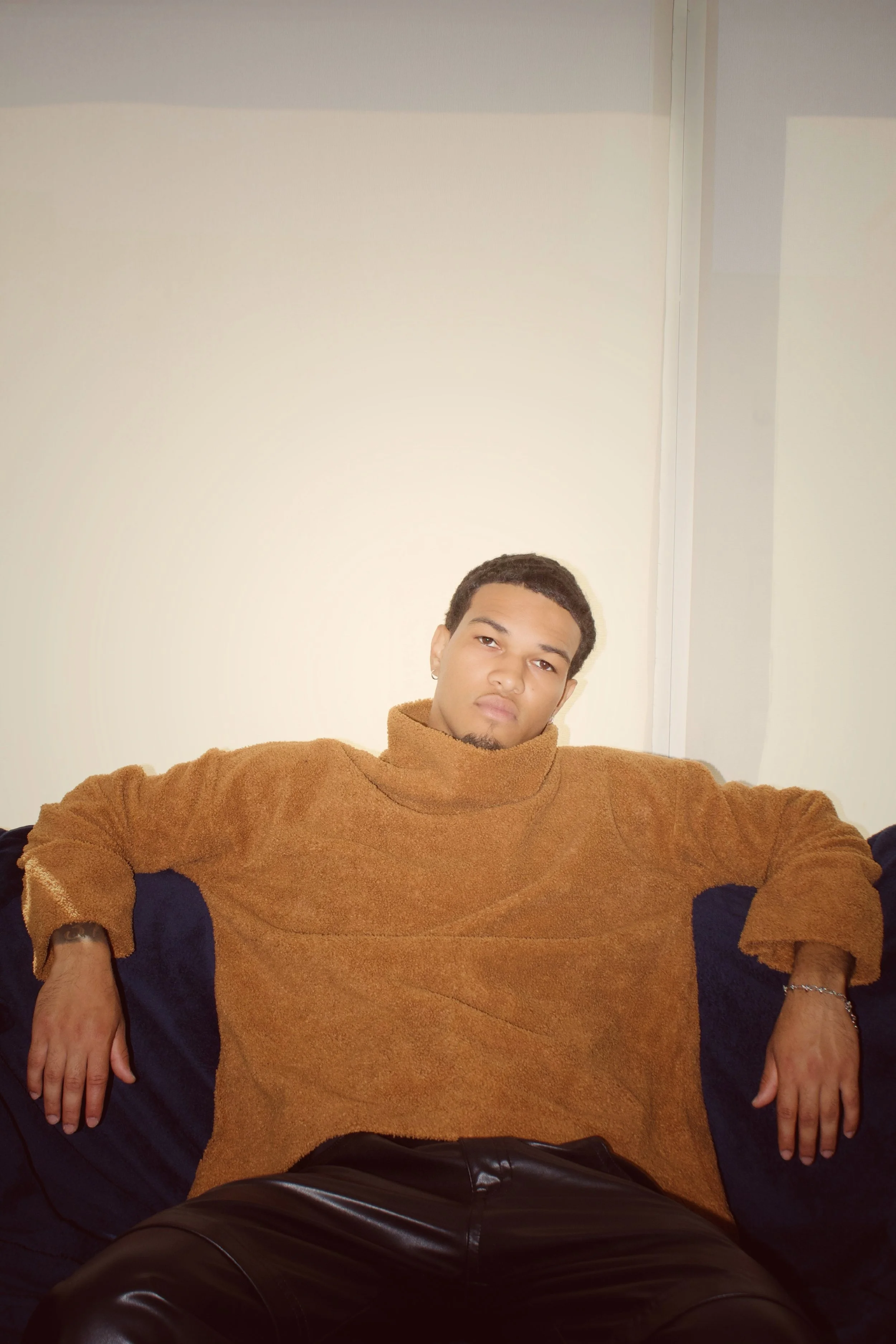 A young man with short curly hair and a serious expression sitting against a plain wall. He is wearing a brown hoodie and black pants.