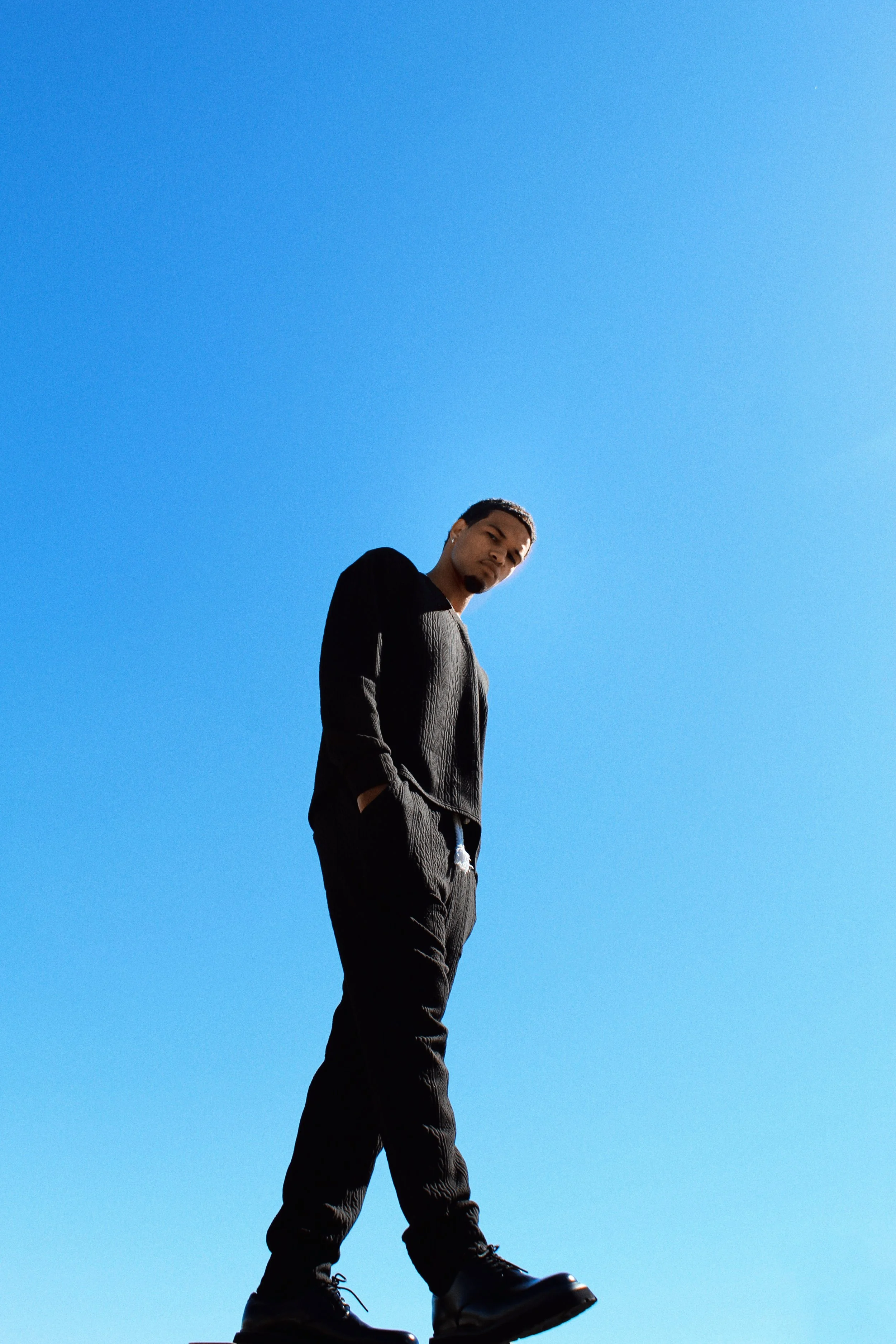 A young man wearing black clothing and boots stands outdoors against a clear blue sky, looking slightly down into the camera.