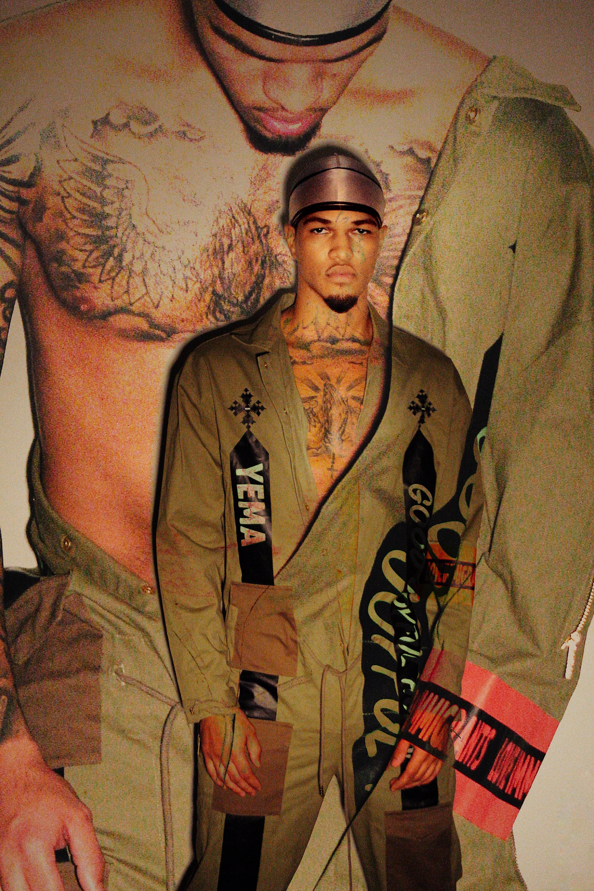 A man with many tattoos, wearing a gray hat and a khaki jacket, standing in front of a poster featuring a close-up of his face and upper body tattoos.