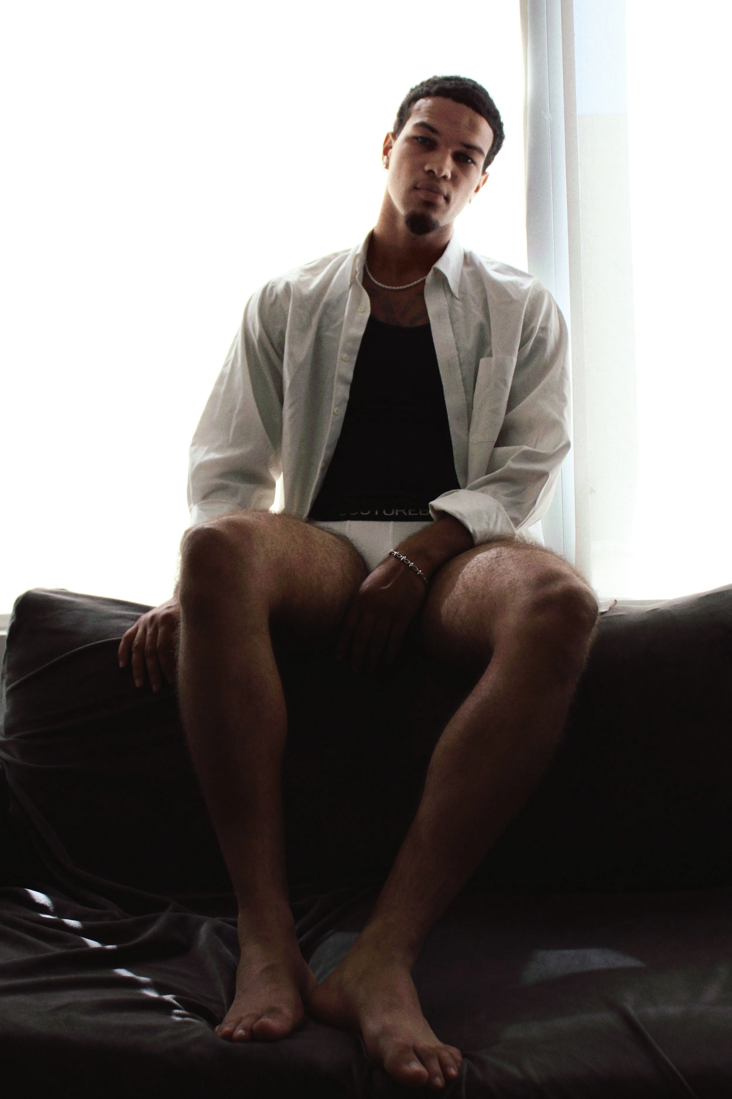 A young man with short dark hair, wearing a white button-up shirt over a black T-shirt, is sitting on a bed with dark bedding near a window with bright light coming through. He has a necklace and a bracelet, and he is looking at the camera with a neu