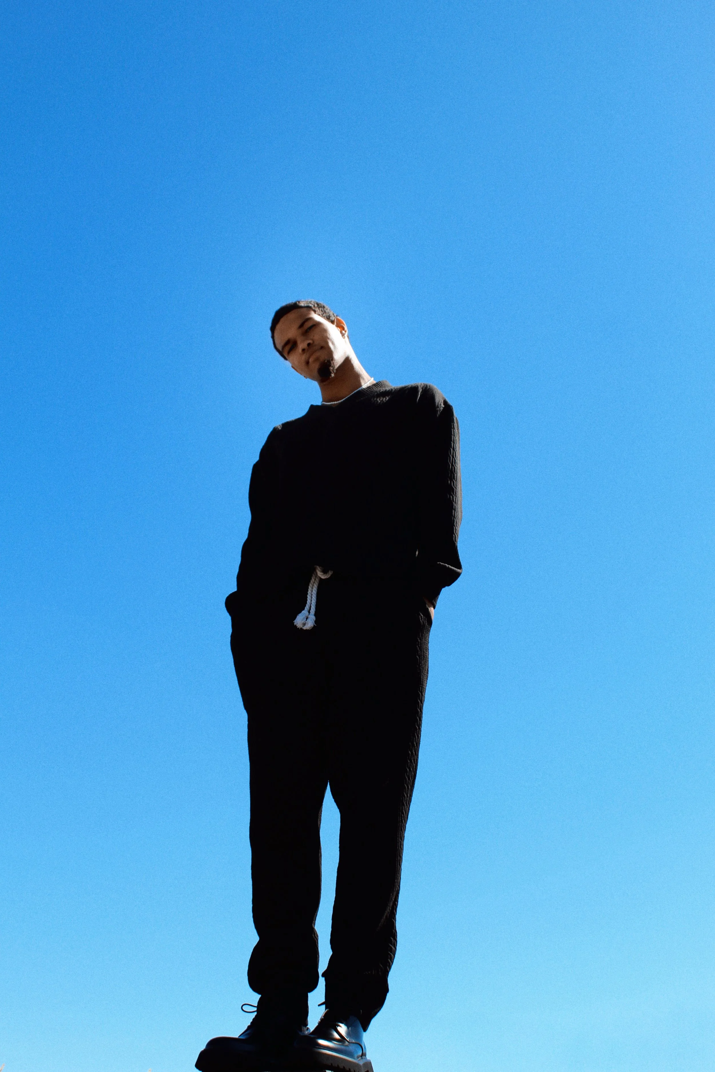A person standing outside wearing black clothing, with arms crossed, against a clear blue sky.
