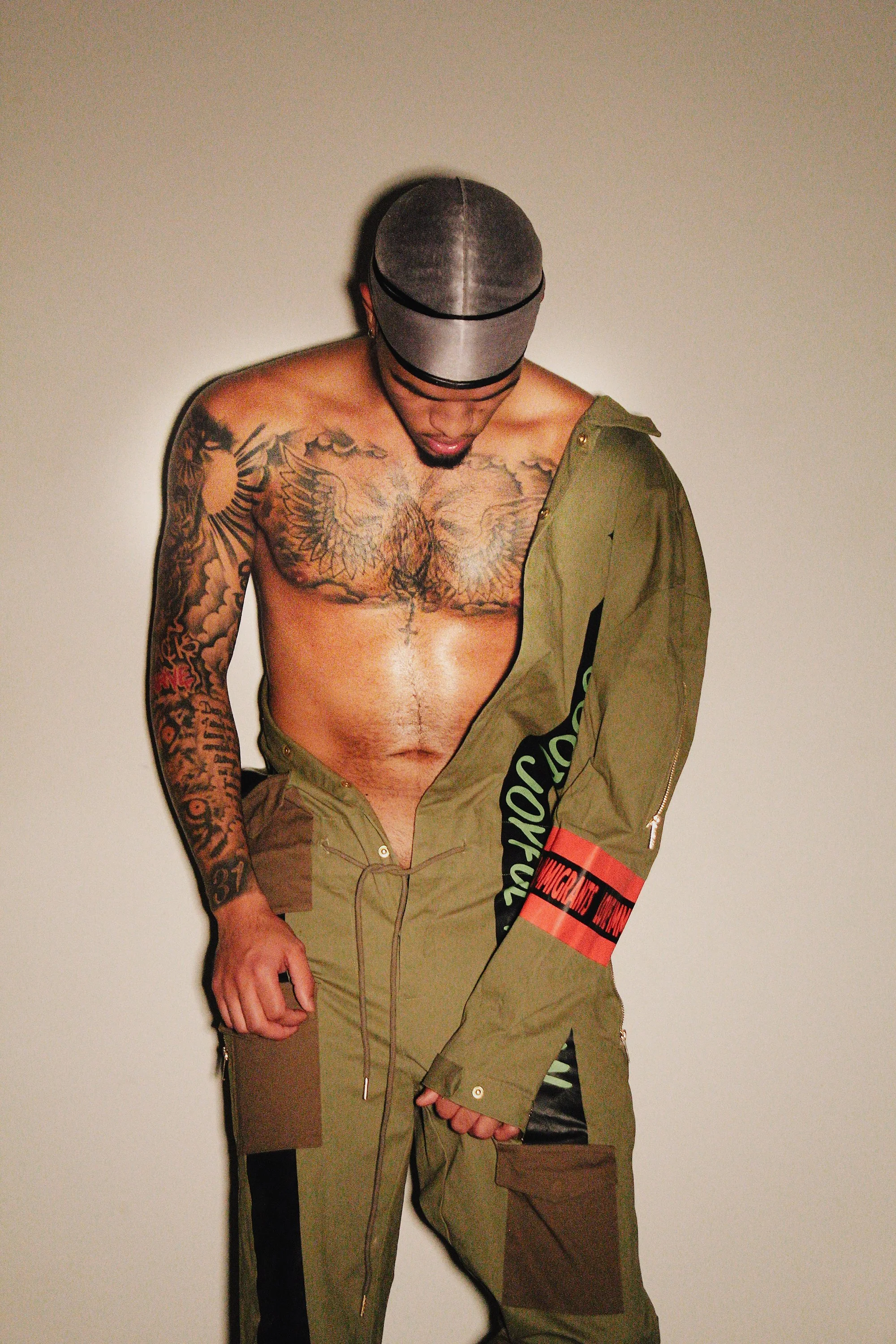 A shirtless man with tattoos on his chest and arms, wearing a black and gray cap, partially dressed in a green jacket and matching green pants against a plain beige background.