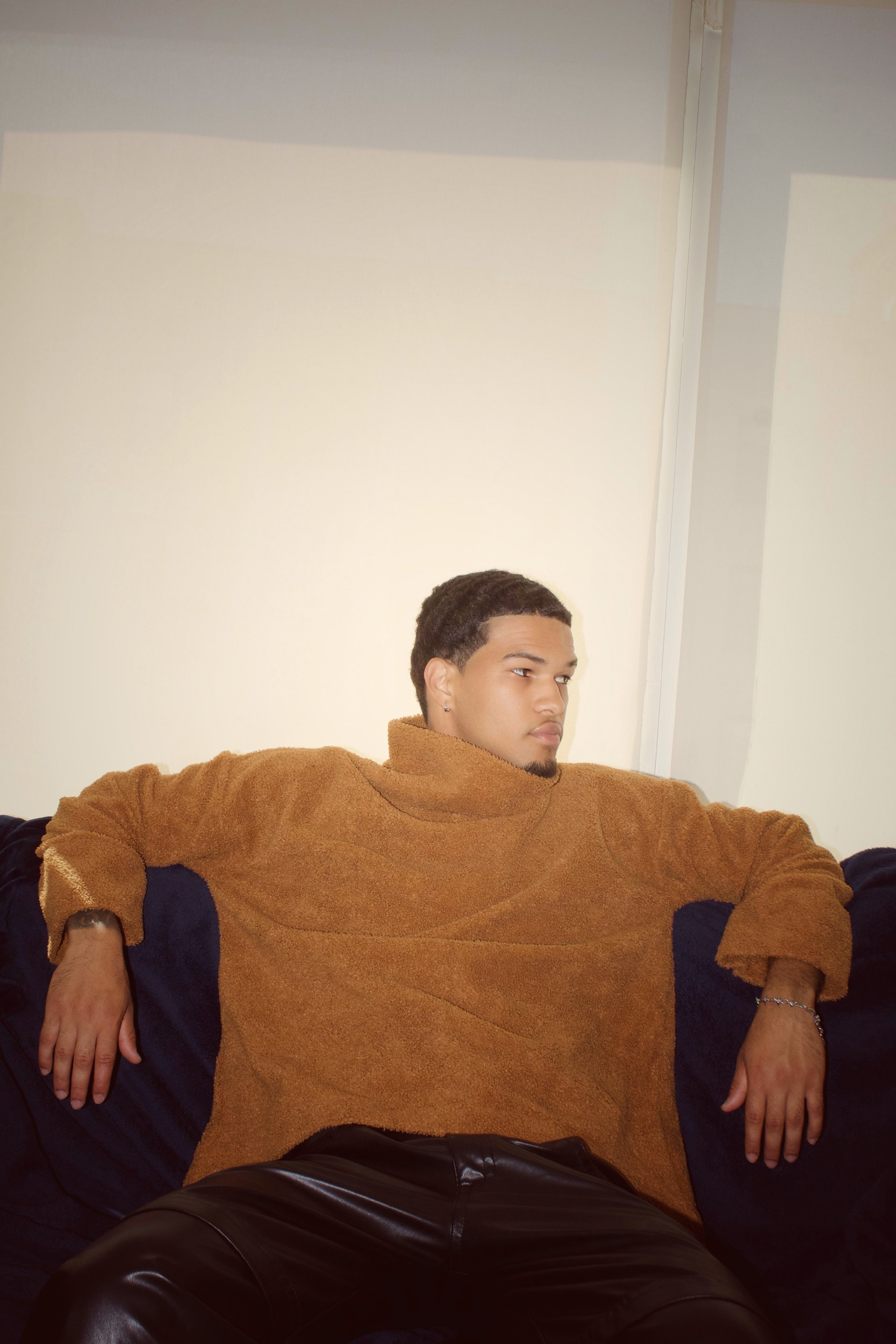 A young man with short dark hair and earrings, wearing a brown sweater, is sitting with his arms resting on the armrests of a black chair. He appears to be in a relaxed pose, looking slightly to his right. The background shows a plain wall and part o
