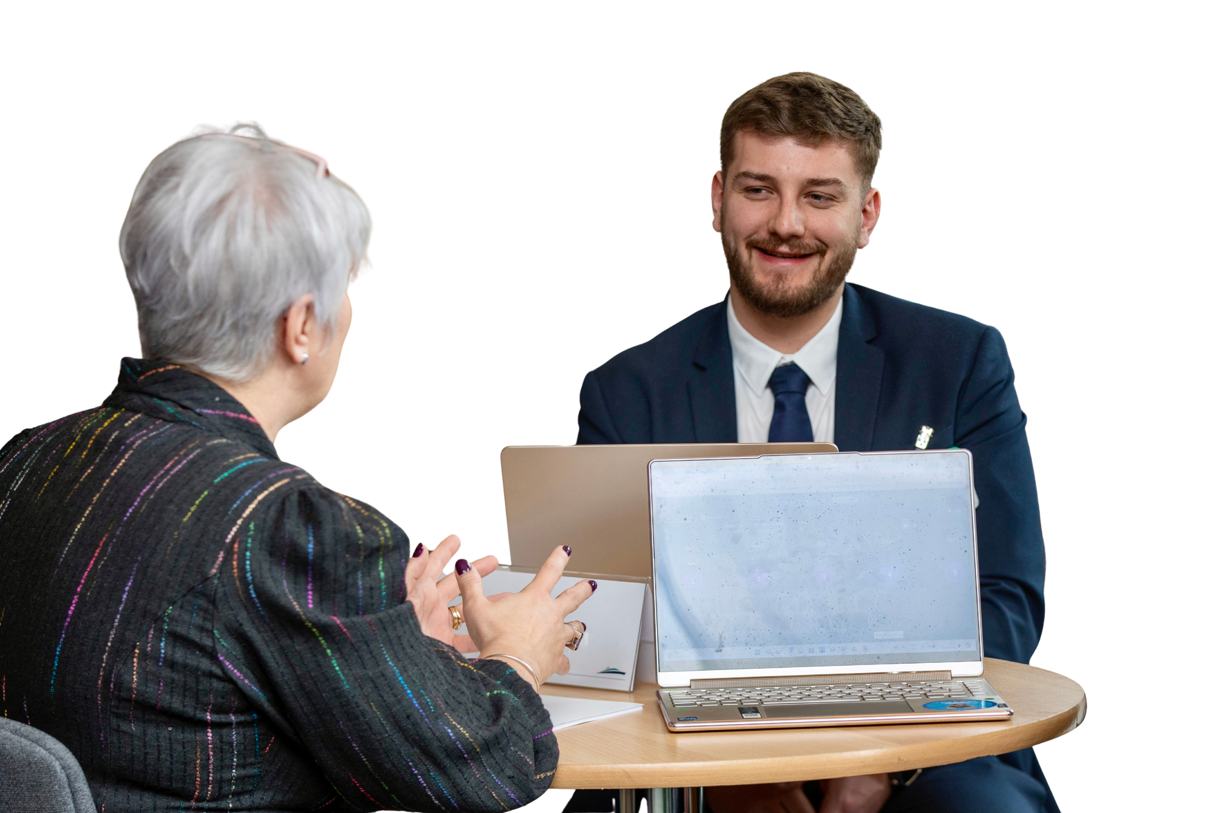 A young man in a suit and tie smiling during a discussion with an older woman with gray hair in a patterned blazer, seated at a round table with laptops.