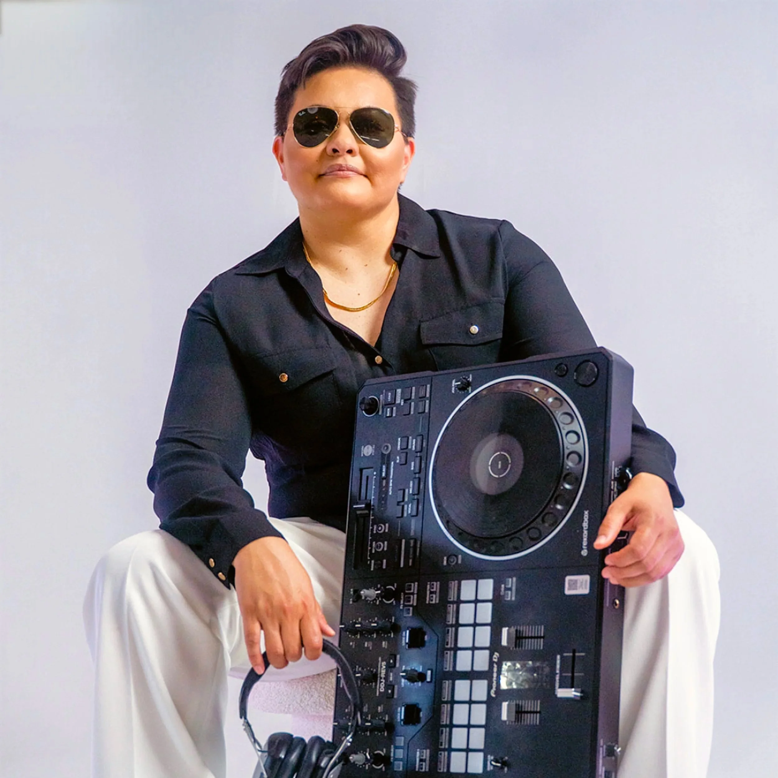 Person wearing sunglasses, black shirt, and white pants sitting with DJ equipment, holding headphones in hand, against a plain white wall.