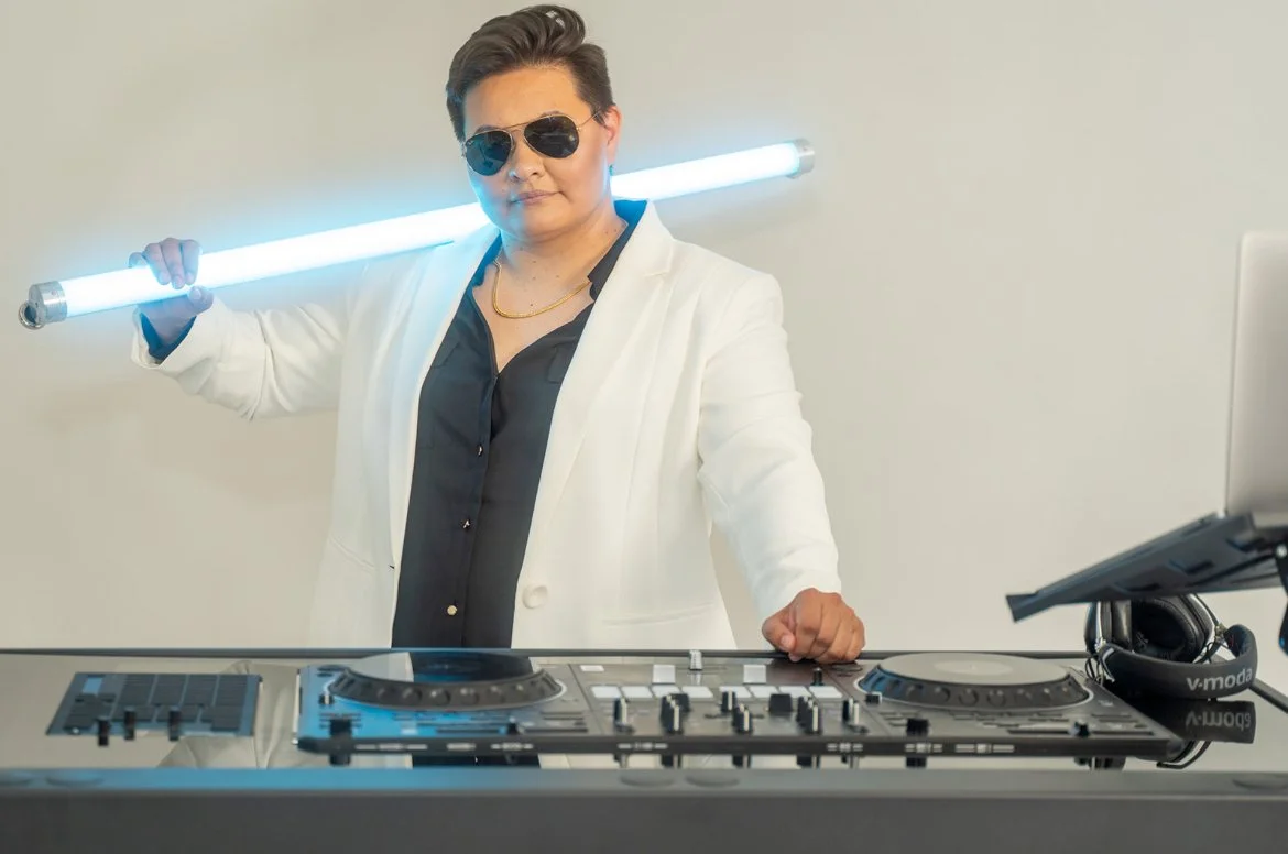 Person wearing sunglasses and a white blazer, holding a glowing blue light tube across their shoulders, standing behind a DJ booth with a laptop and headphones.