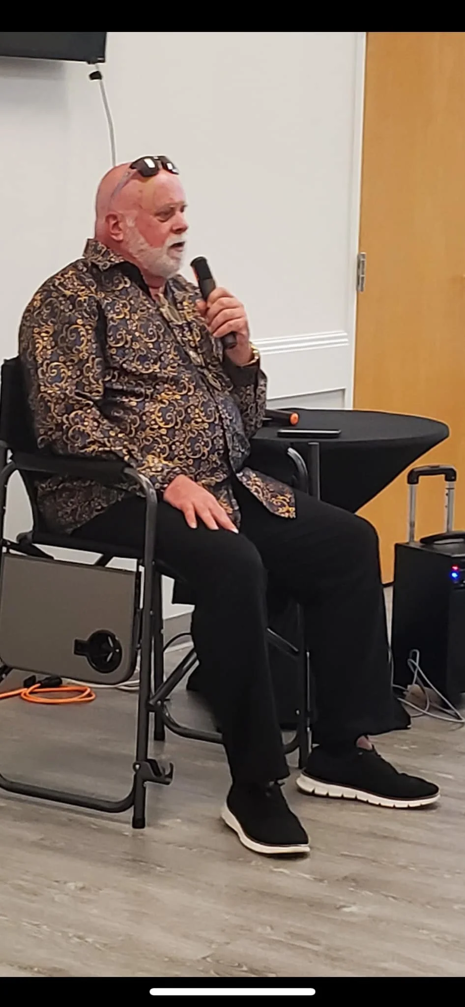 An elderly man with a white beard, seated in a wheelchair, holding a microphone and speaking. He is wearing a patterned shirt and black pants and has sunglasses resting on his bald head. A small table with a speaker, a mobile phone, and some cords are nearby, suggesting he is giving a speech or presentation in an indoor setting.