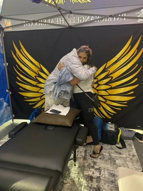 Two women hugging in front of a black backdrop with large yellow wing designs, standing behind a massage table in an indoor setting.