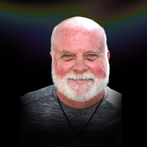 Close-up of a smiling older man with a white beard and bald head, wearing a gray shirt and a black necklace, against a dark background.