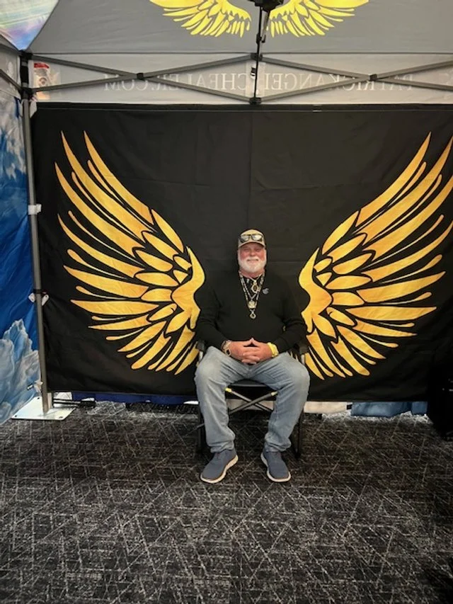 A man sitting in front of a black backdrop with large yellow wings and a yellow winged emblem above him at a booth or event.