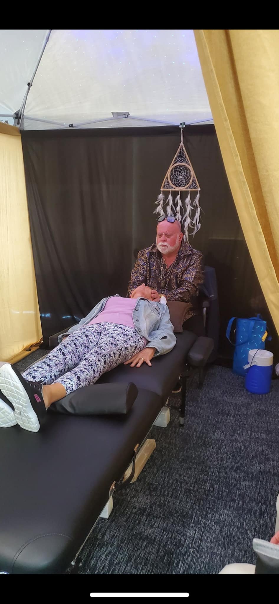 A person lies on an examination or massage table in a tent, with a man sitting and holding their hand. The tent has black and yellow walls, a dreamcatcher hanging above, and a starry sky projected on the ceiling.