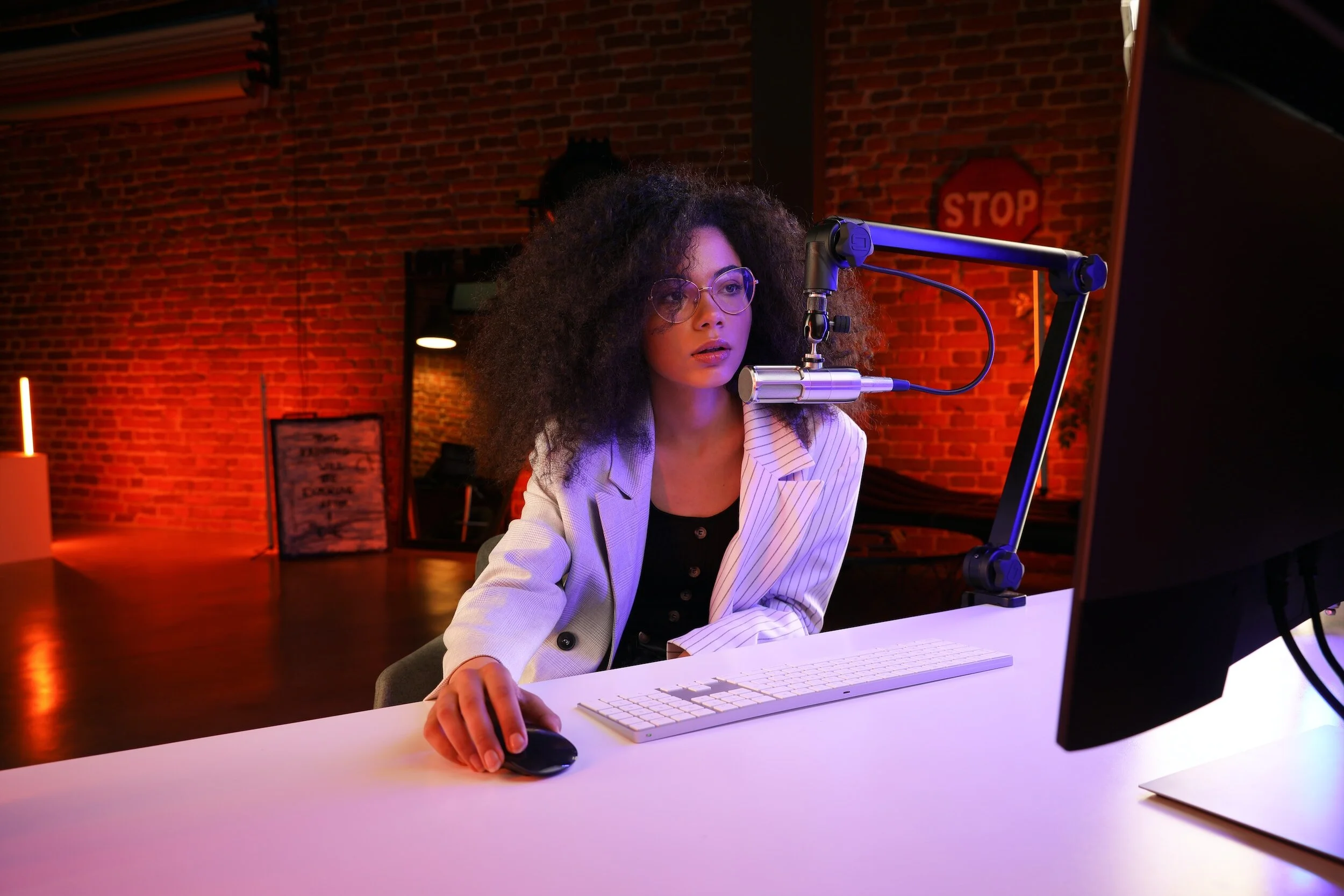 Person in a striped blazer using a computer with a microphone setup, sitting in a room with brick walls and soft lighting.