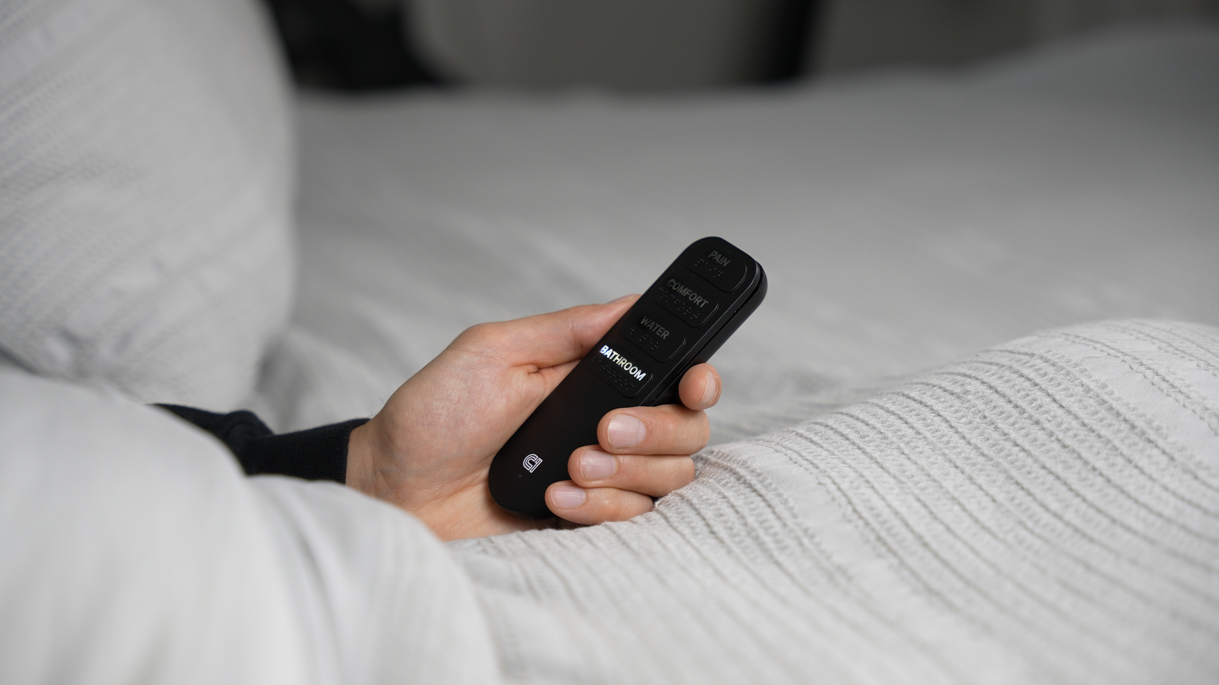 Person holding a remote control with buttons labeled 'PAIN,' 'COMFORT,' 'WATER,' and 'BATHROOM,' resting on a bed with white bedding.