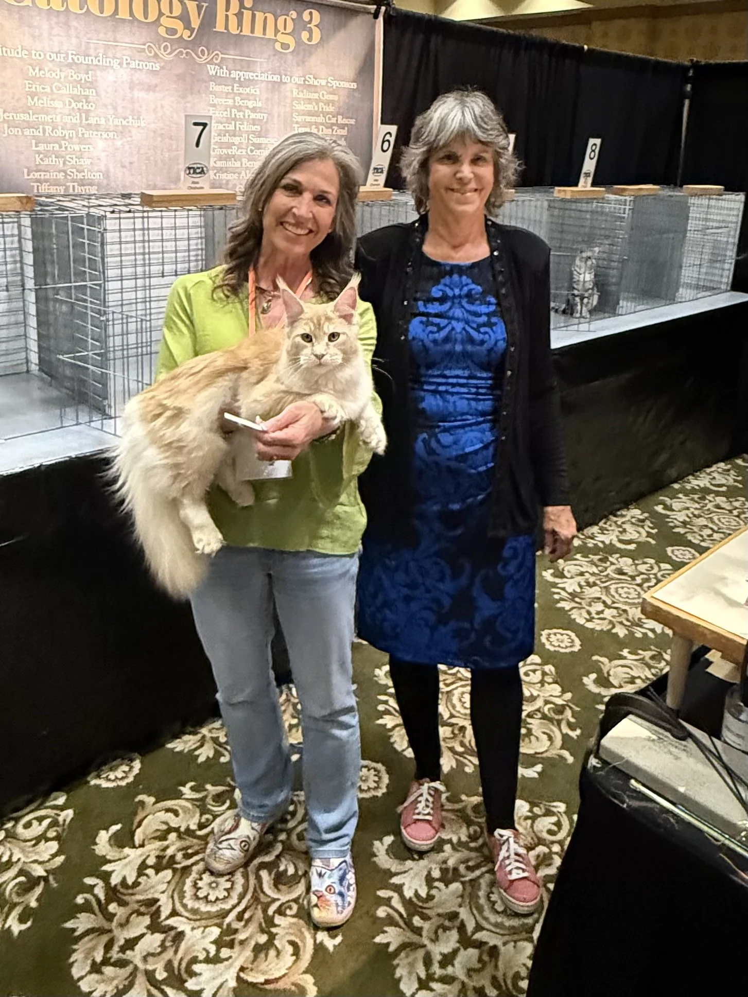 Two women standing indoors at a cat show, with cages in the background. The woman on the left holds a large, fluffy orange and white cat, both smiling at the camera. The woman on the right wears a blue dress with a black jacket. The floor has a patte