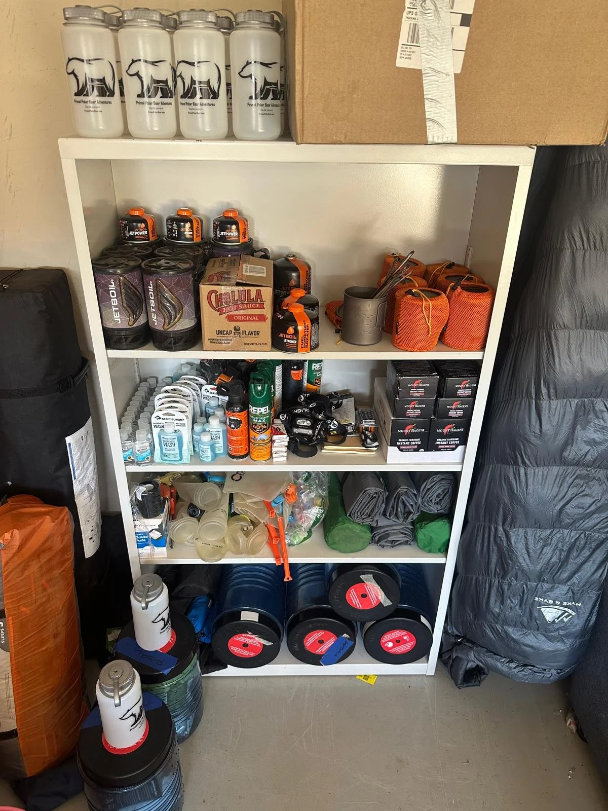 A white shelving unit holding water bottles, boxes, cans, sports gear, and camping supplies.