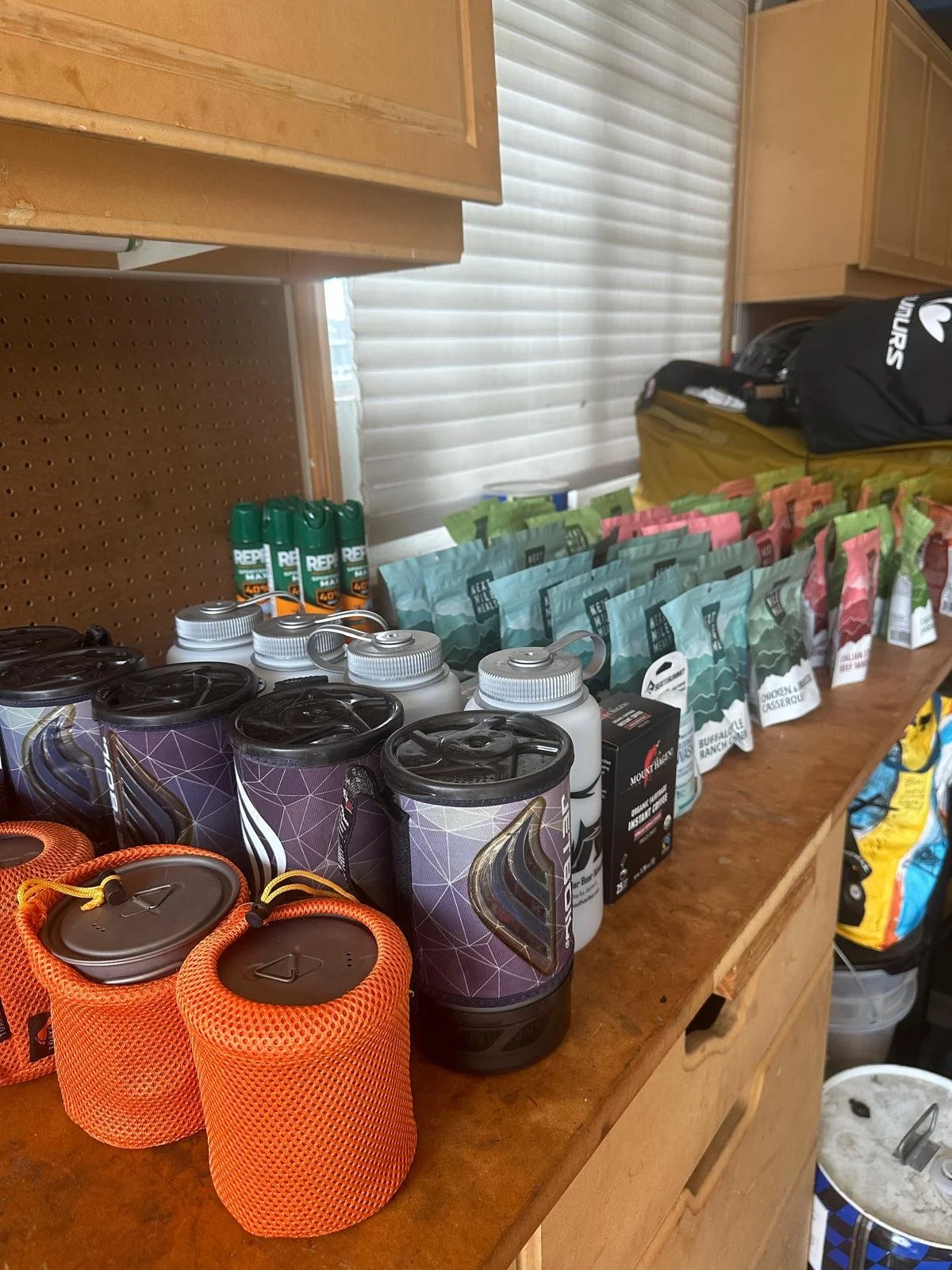 Assorted packaged snacks, drinks, and supplies arranged on a wooden table, with a window and blinds in the background.