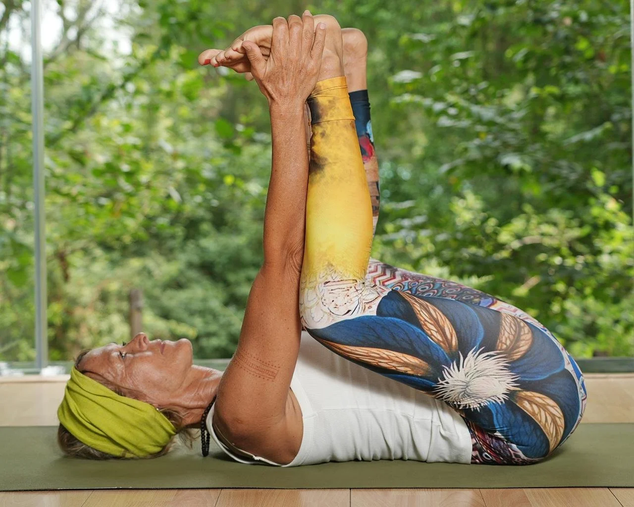 Ältere Frau macht eine Yoga-Übung auf einer Matte, trägt bunte Leggings, ein weißes Top und ein grünes Stirnband, vor einer großen Fensterfront mit Blick auf grüne Bäume.