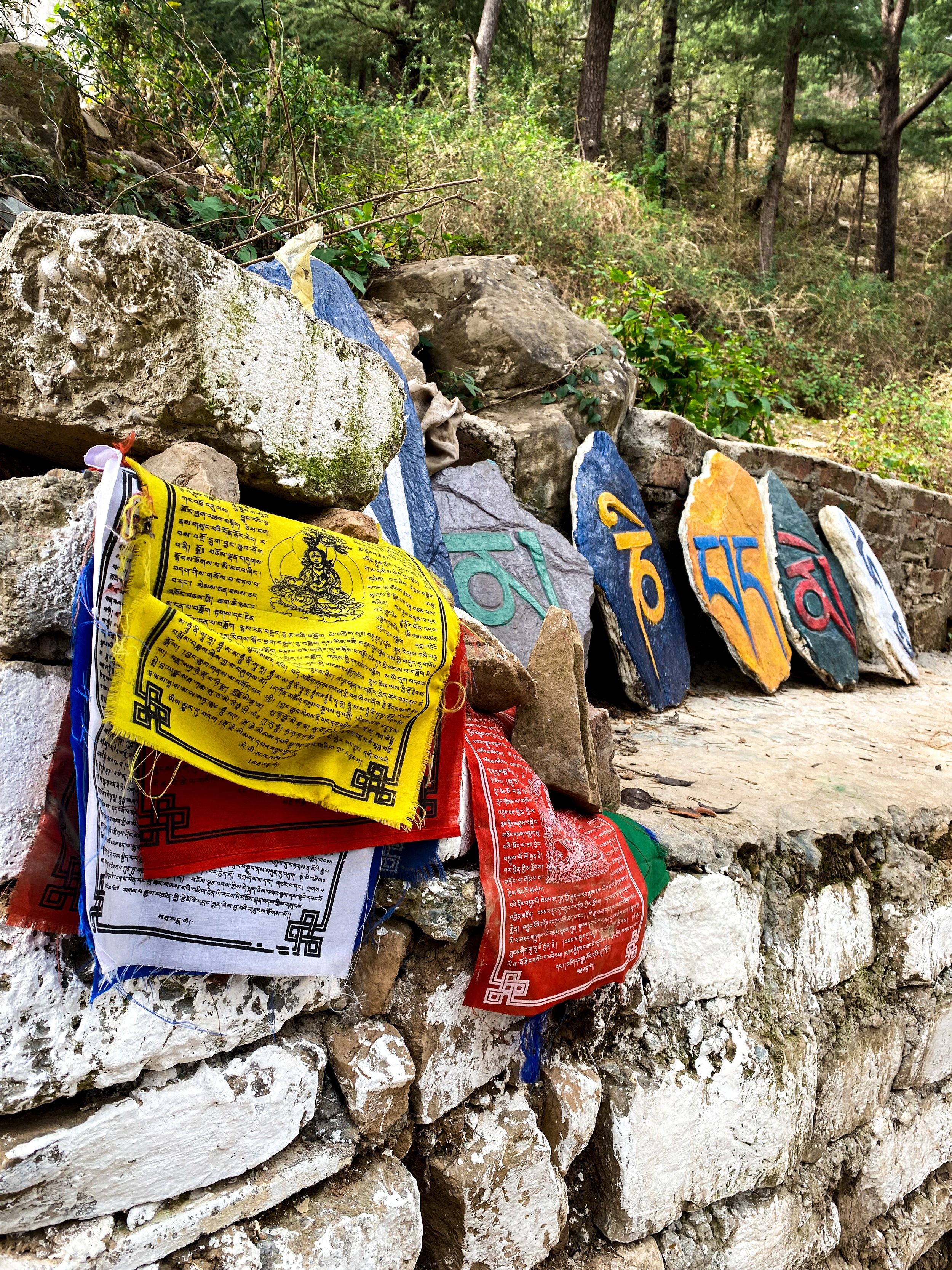 Bunte Opfergaben und Gebetsfahnen liegen auf einer steinernen Mauer in einer bewaldeten Umgebung.