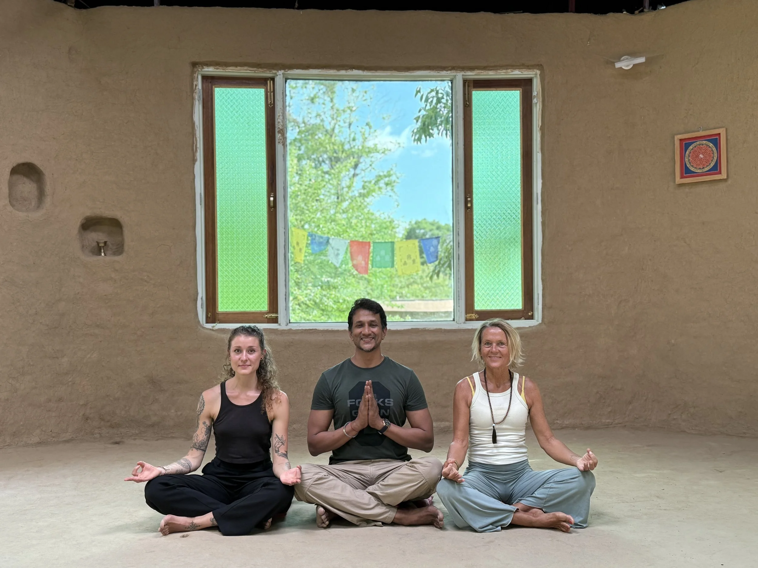 Drei Menschen in Meditationshaltung sitzen im Raum vor einem großen Fenster mit grünen, gemusterten Fensterläden. Draußen sind Bäume und blauer Himmel sichtbar. Der Raum hat eine unverputzte Wand mit kleinen Nischen, in denen Kerzen stehen. An der Wand hängt ein farbenfrohes Bild.