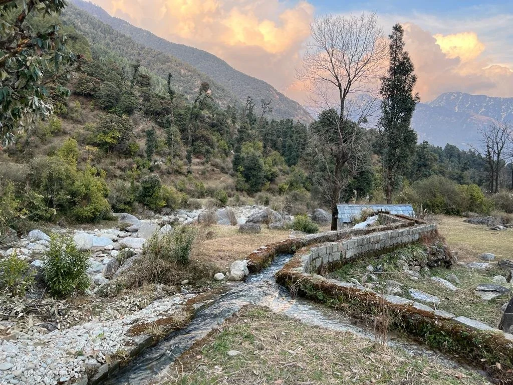 Ein bewaldetes Tal mit einem kleinen Bach, der in eine künstliche Wasserleitung mündet, umgeben von Bergen und Bäumen, bei Sonnenuntergang.