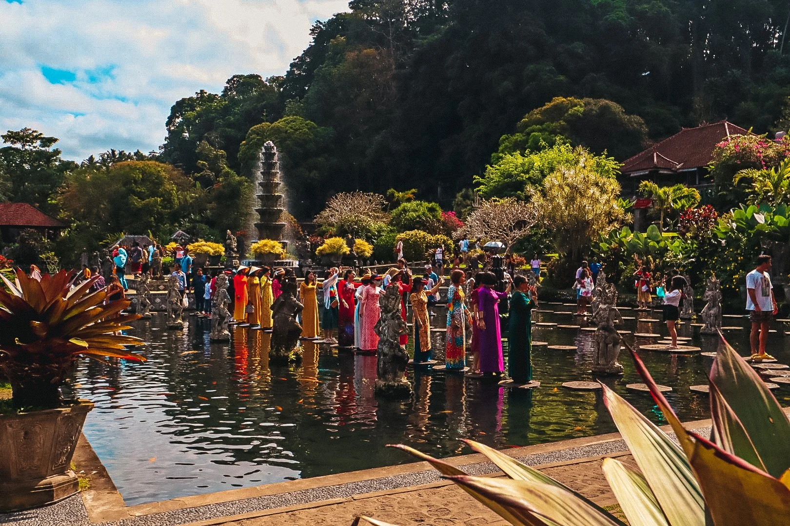 Menschen in traditionellen bunten Kleidern, die in einem Wasserbecken stehen, umgeben von Skulpturen und Pflanzen, in einem japanischen Garten mit Hügel und Wasserfontäne im Hintergrund.