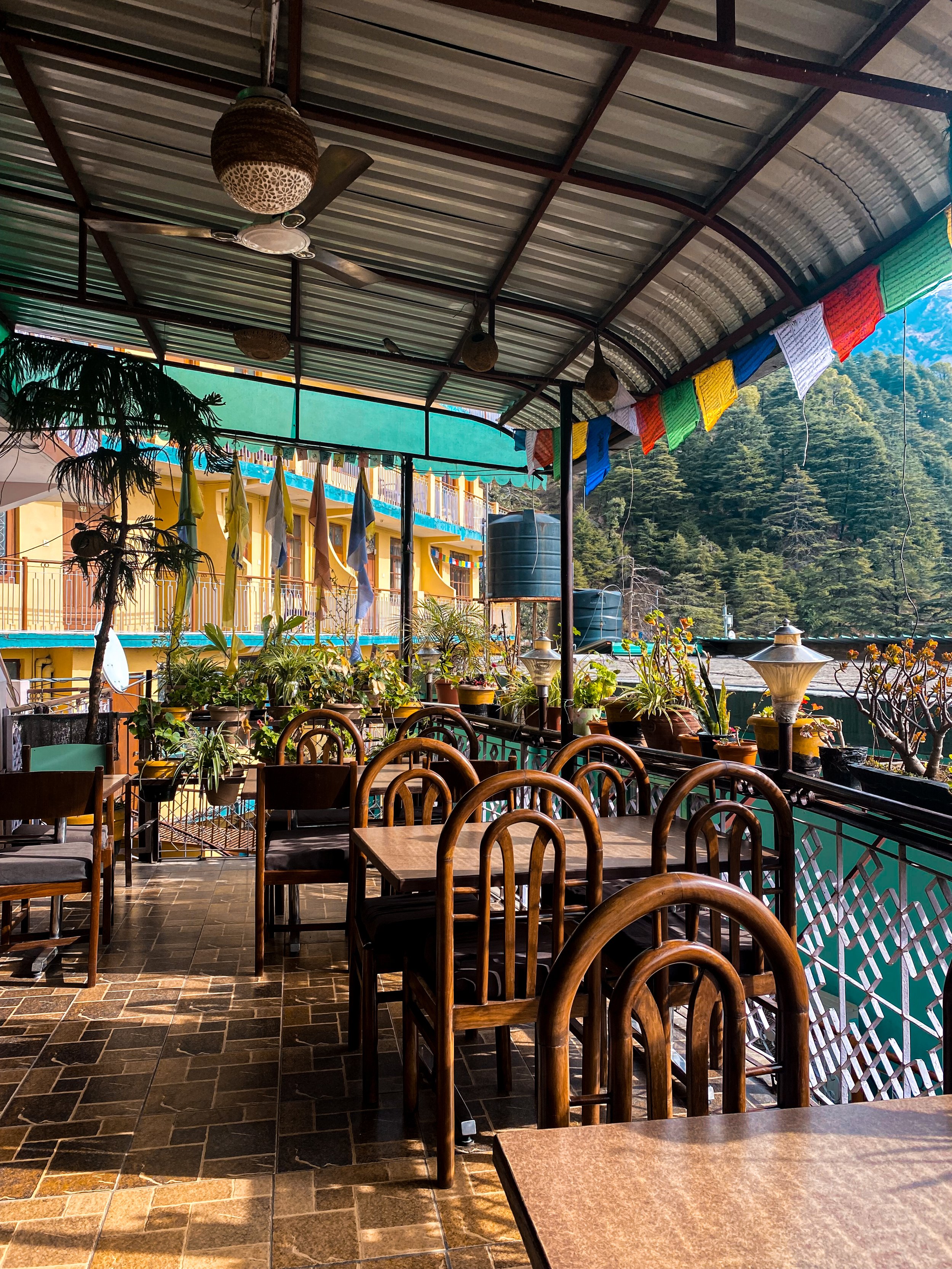 Balkon mit Holztischen und Stühlen, viele Topfpflanzen, bunte Gebetsflaggen, Wasserbehälter, mit Blick auf Häuser und Berge im Hintergrund.