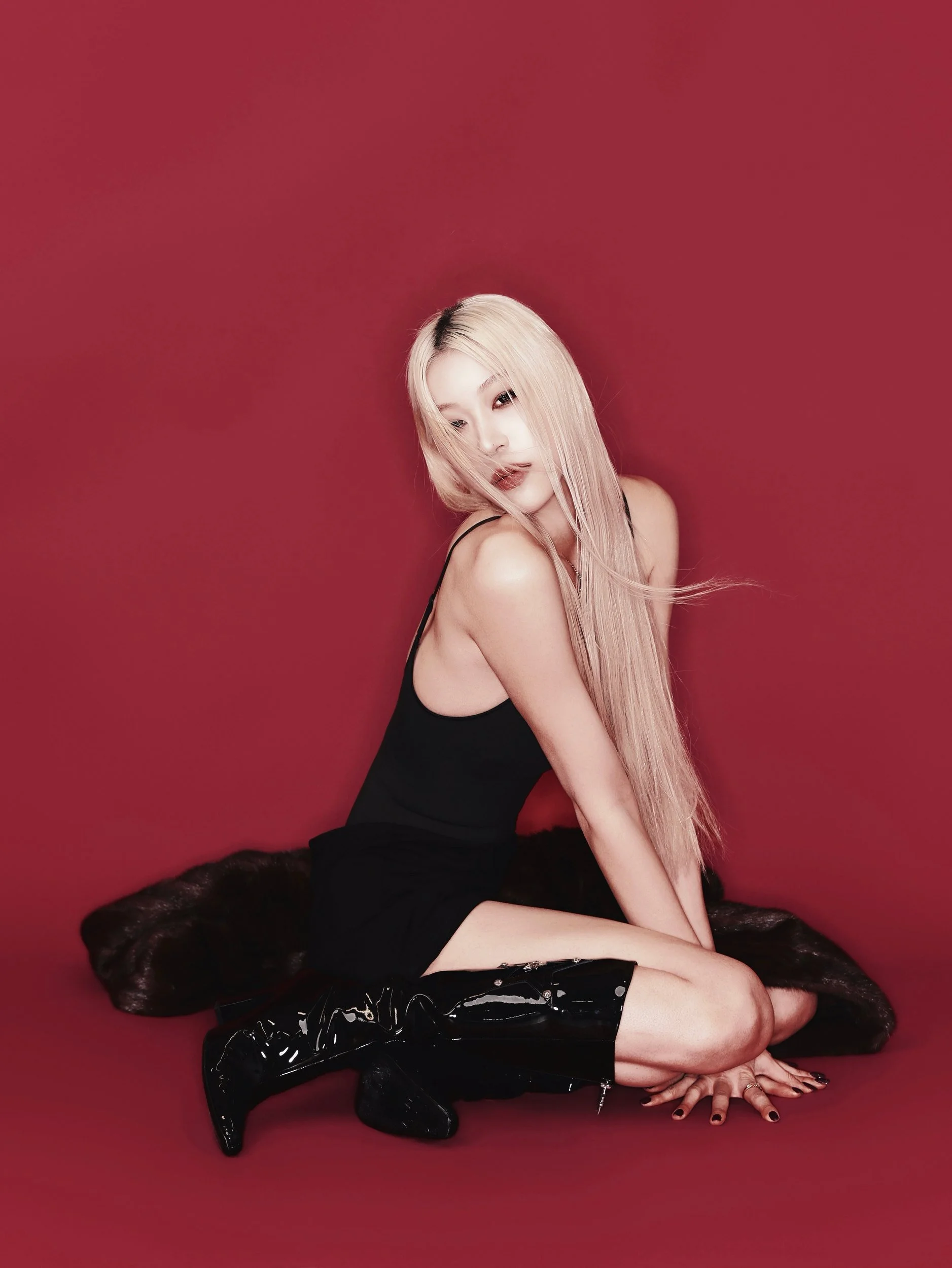 Full-length studio portrait of a woman seated in boots on a red backdrop