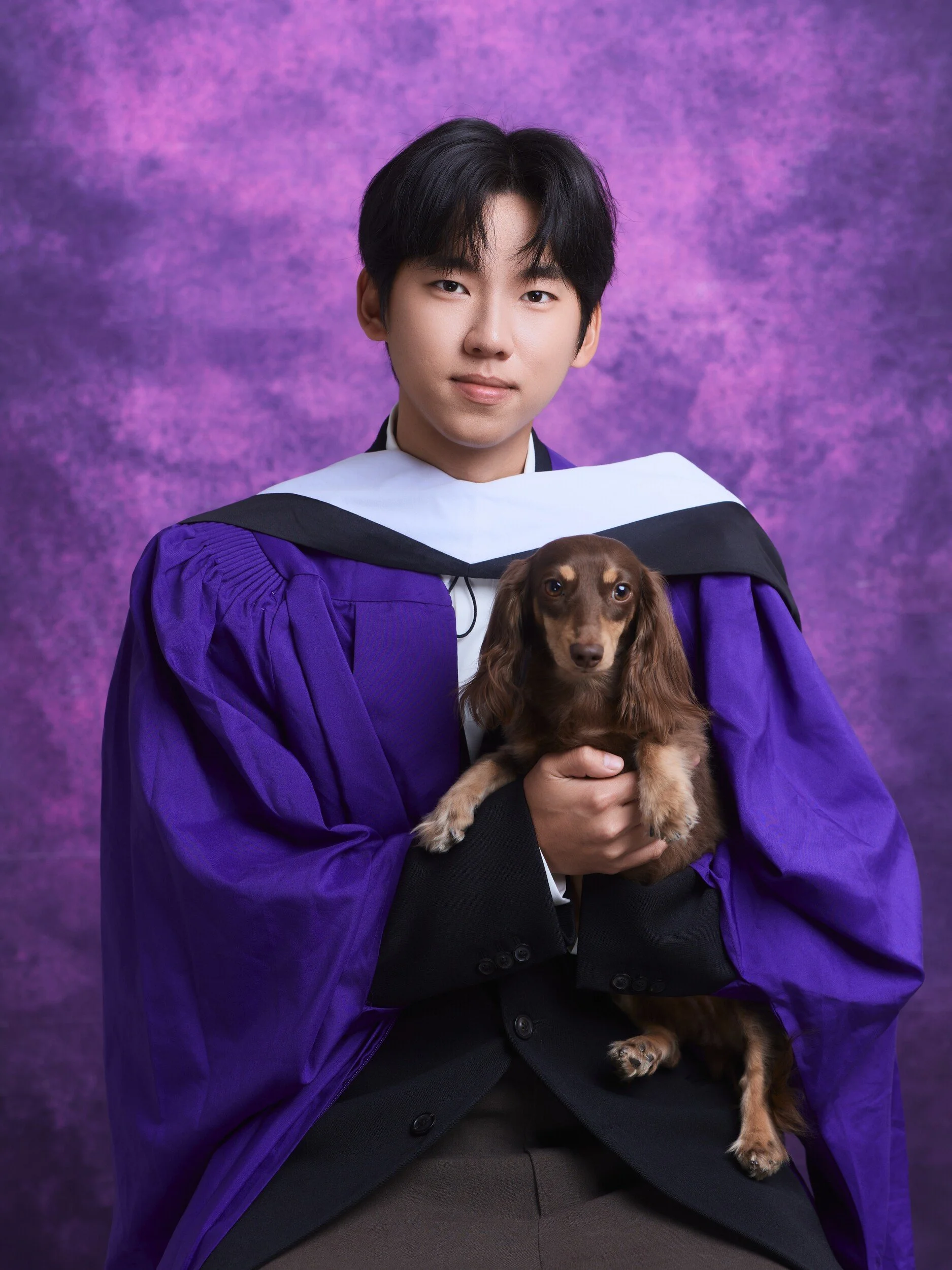 Graduation portrait of a young man in a purple gown holding his dachshund