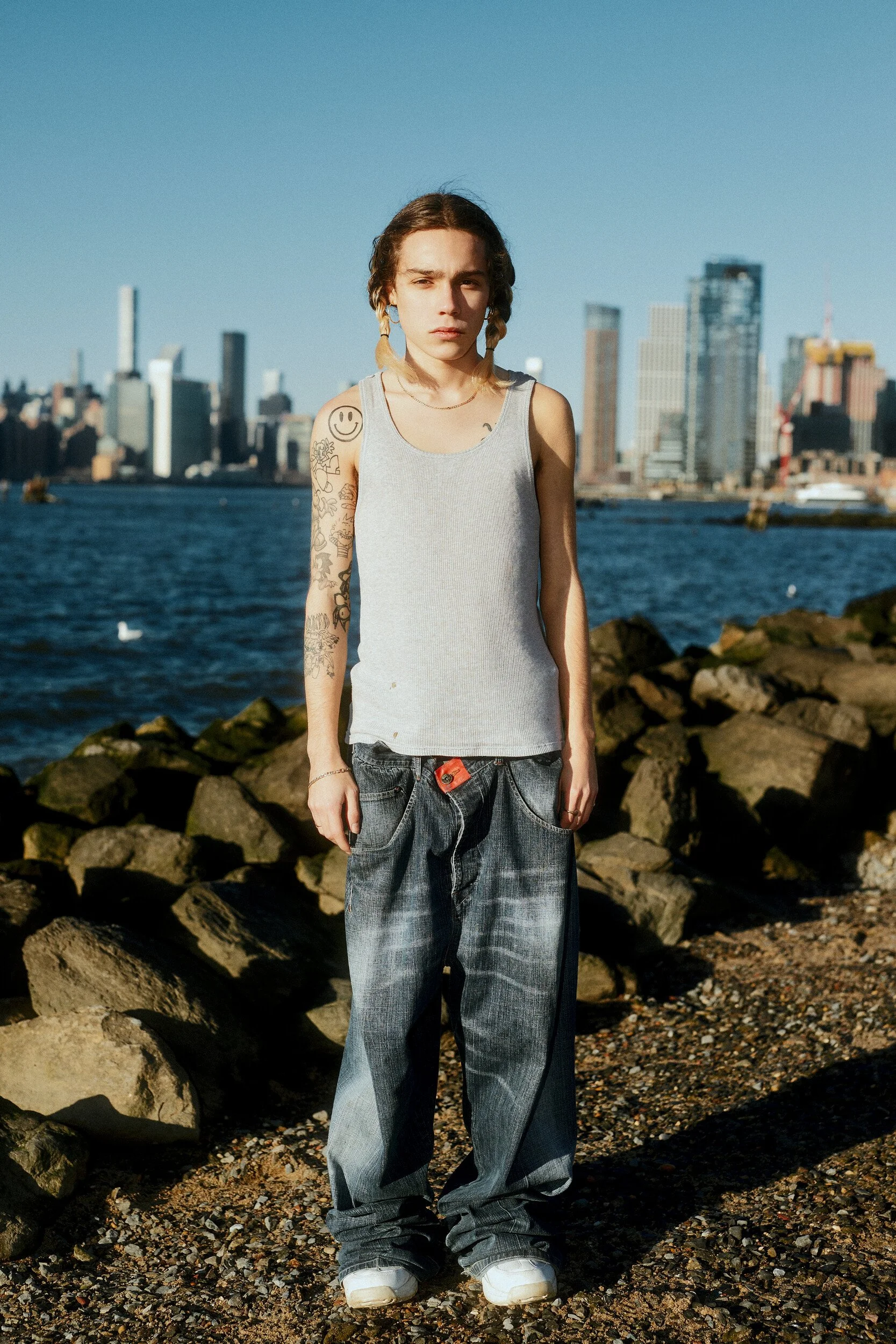 Model digitals by the river: full-length front view of a tattooed man in a grey tank top and loose jeans