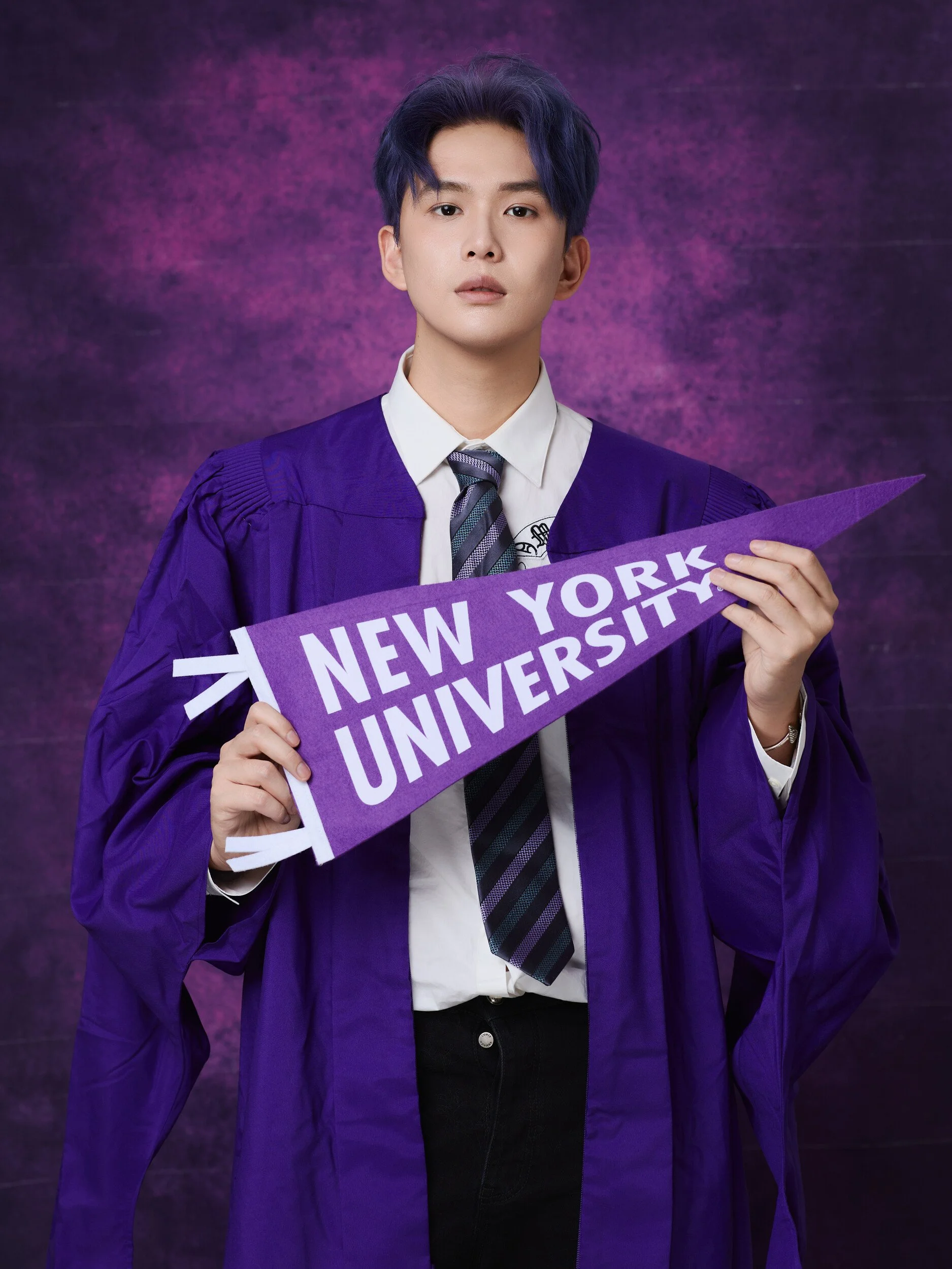NYU graduation portrait of a young man with purple hair holding a New York University pennant