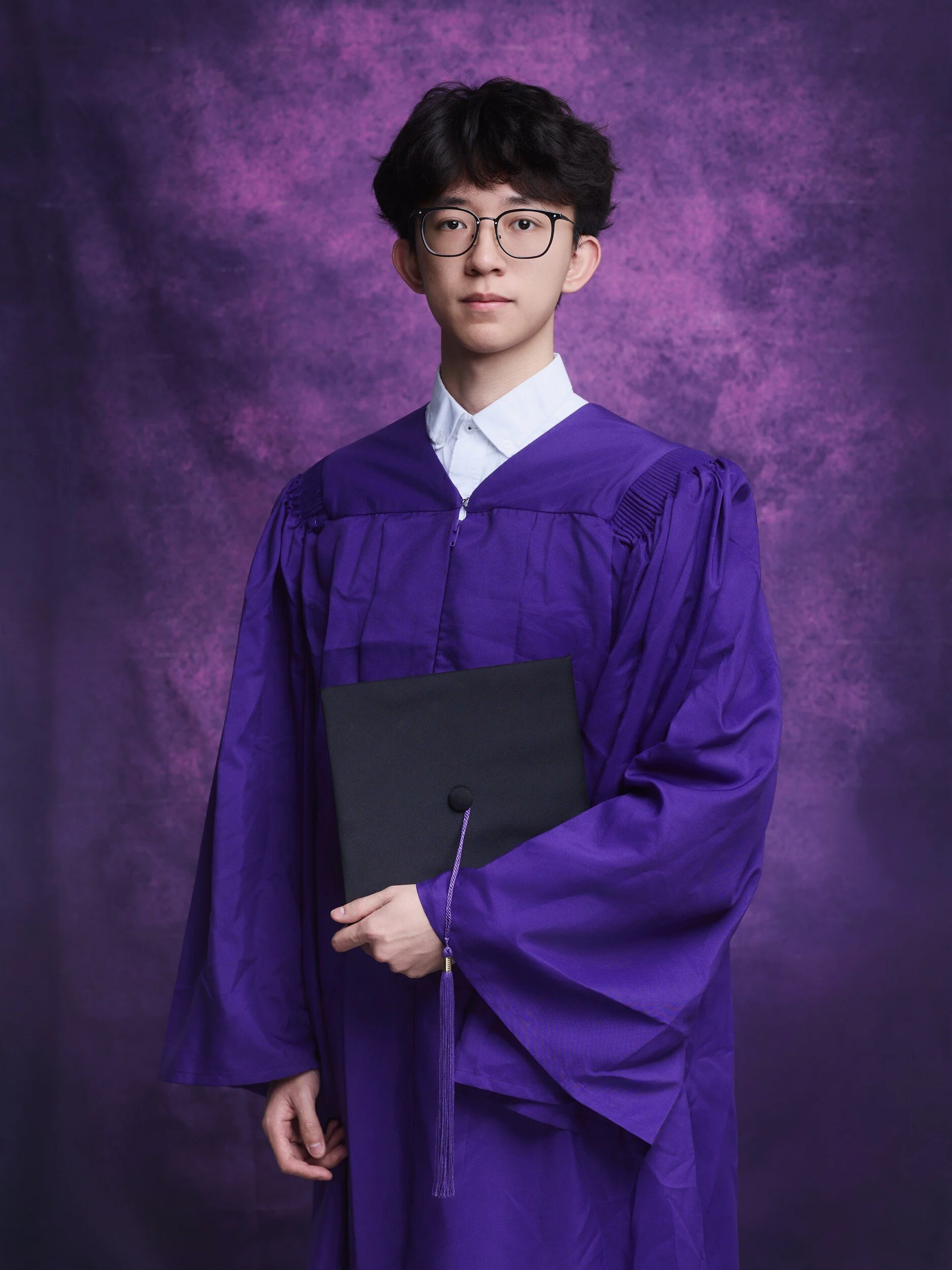 Full-length NYU graduation portrait of a young man in glasses holding his cap