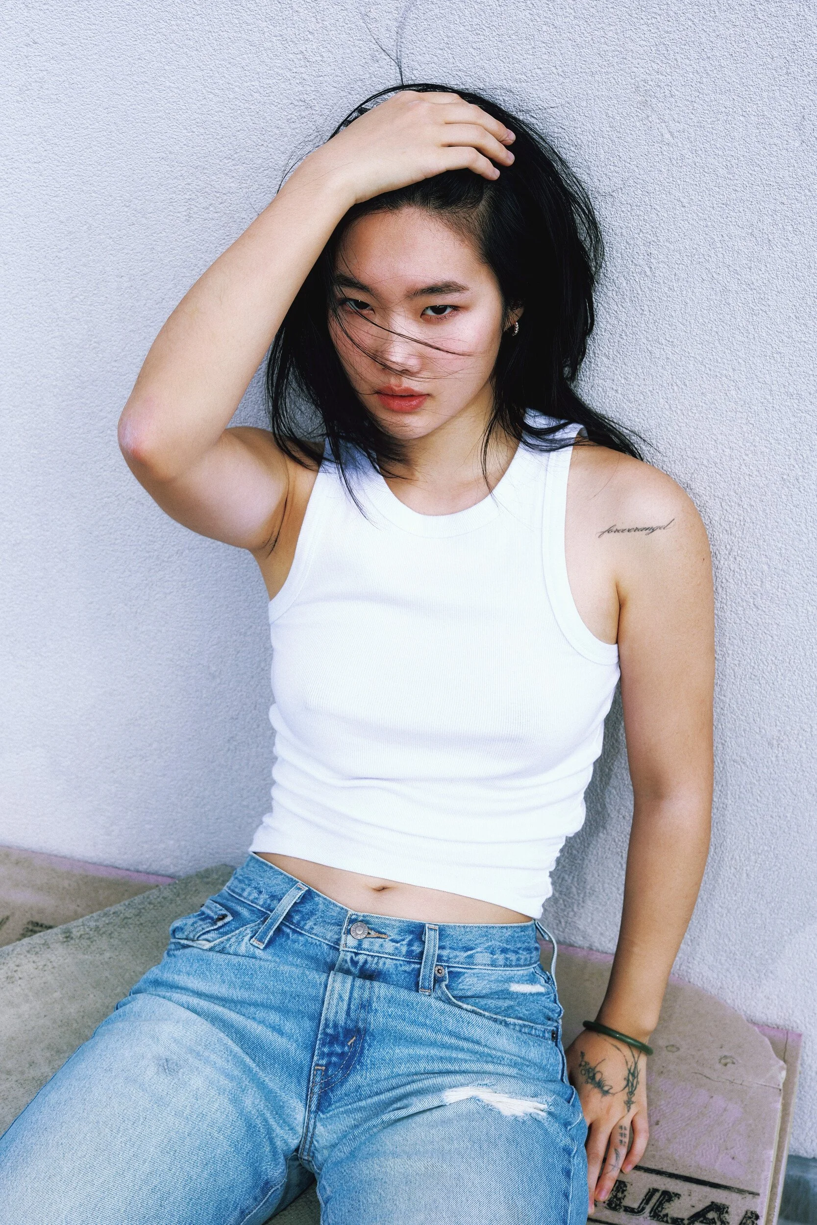 Model digitals outdoors: young woman in a white tank top and jeans with one hand raised to her hair, seated against a sunlit wall