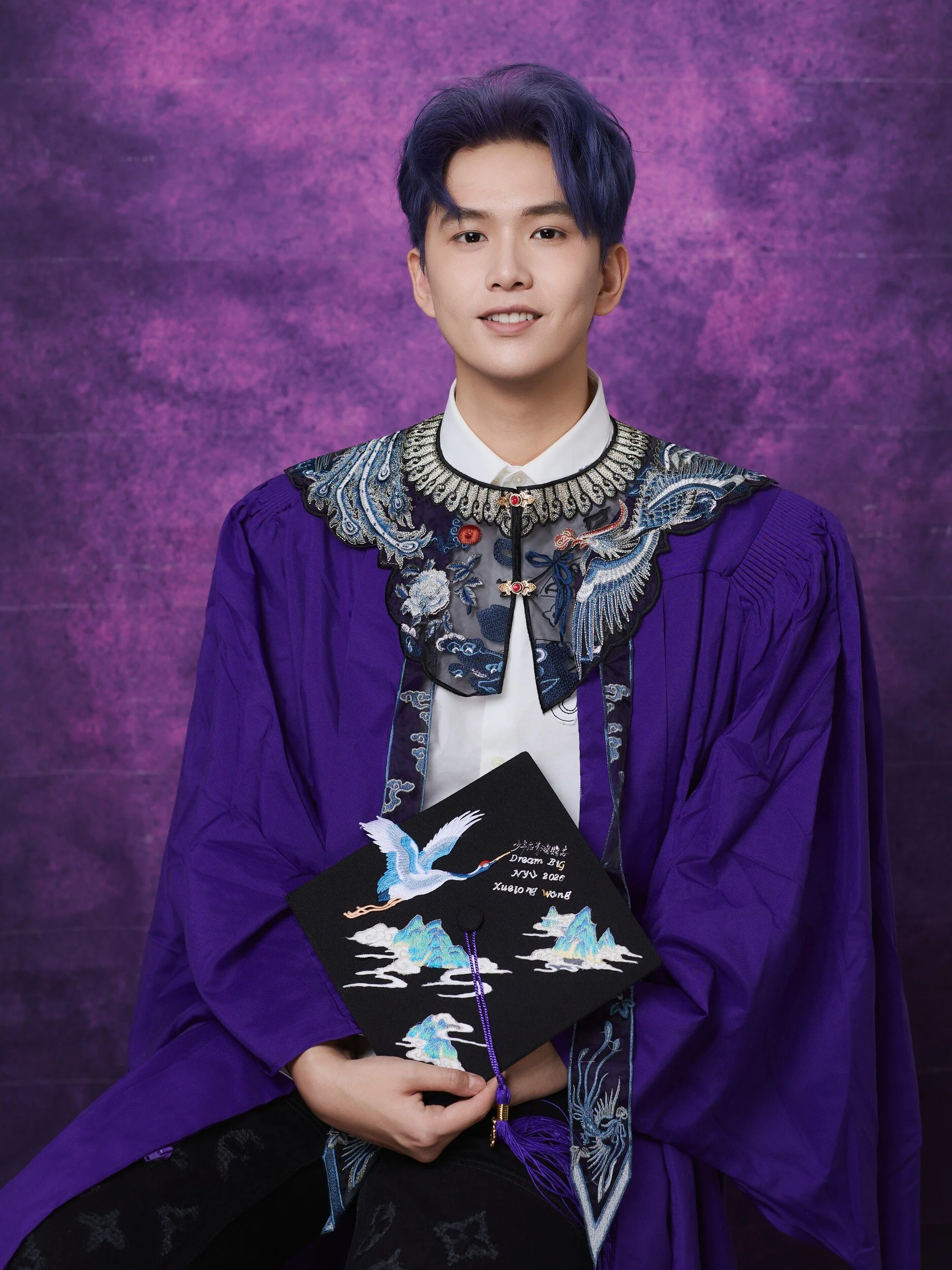 Seated graduate with blue-toned hair holding a hand-painted mortarboard