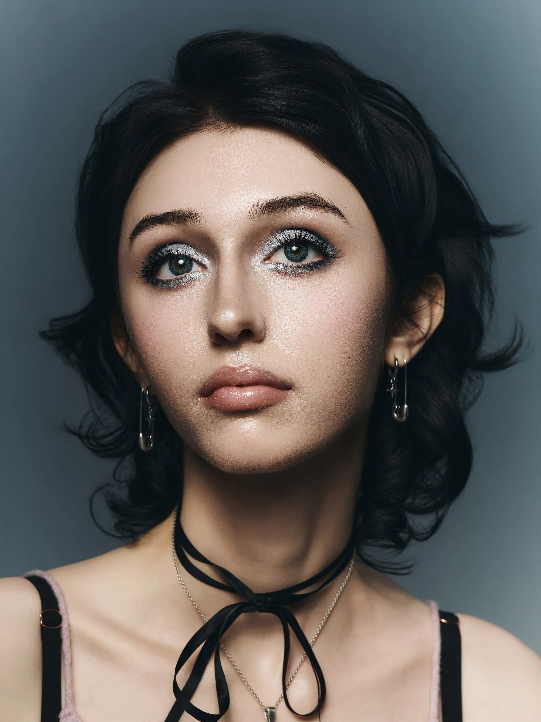 Beauty portrait of a woman with dark curls, silver-blue shimmer eyes and nude lips, black ribbon choker on a slate backdrop
