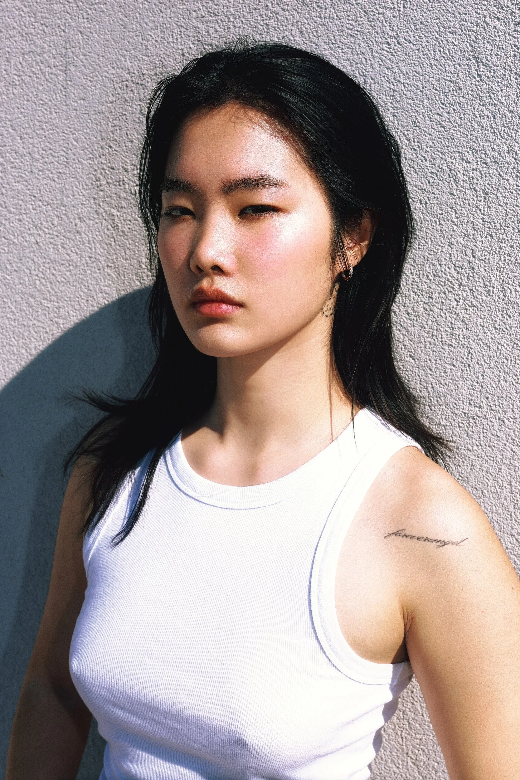 Model digitals outdoors: close-up of a young woman in a white tank top looking toward the camera, dark shoulder-length hair, sunlit wall