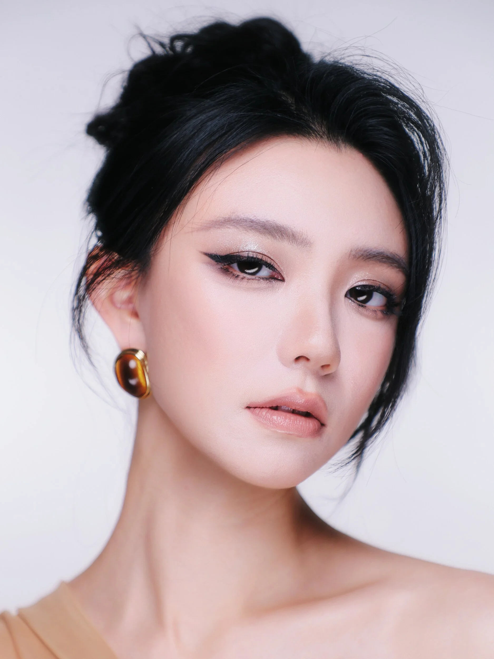 Three-quarter beauty portrait of a woman with a soft updo, winged liner and amber hoop earrings on a pale lilac backdrop