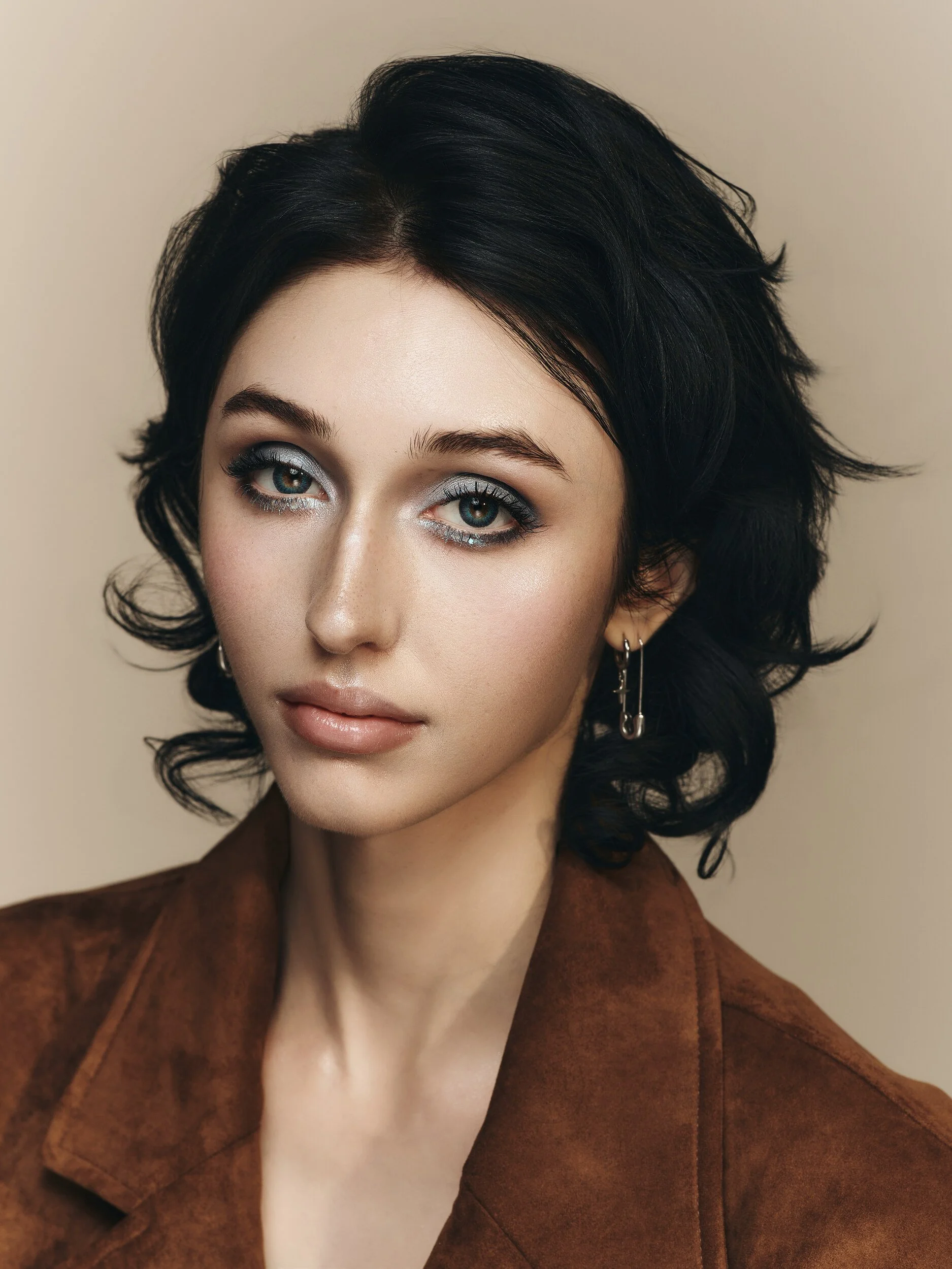 Front-facing beauty portrait with shimmering eye makeup, glossy skin and safety-pin earrings on a beige backdrop