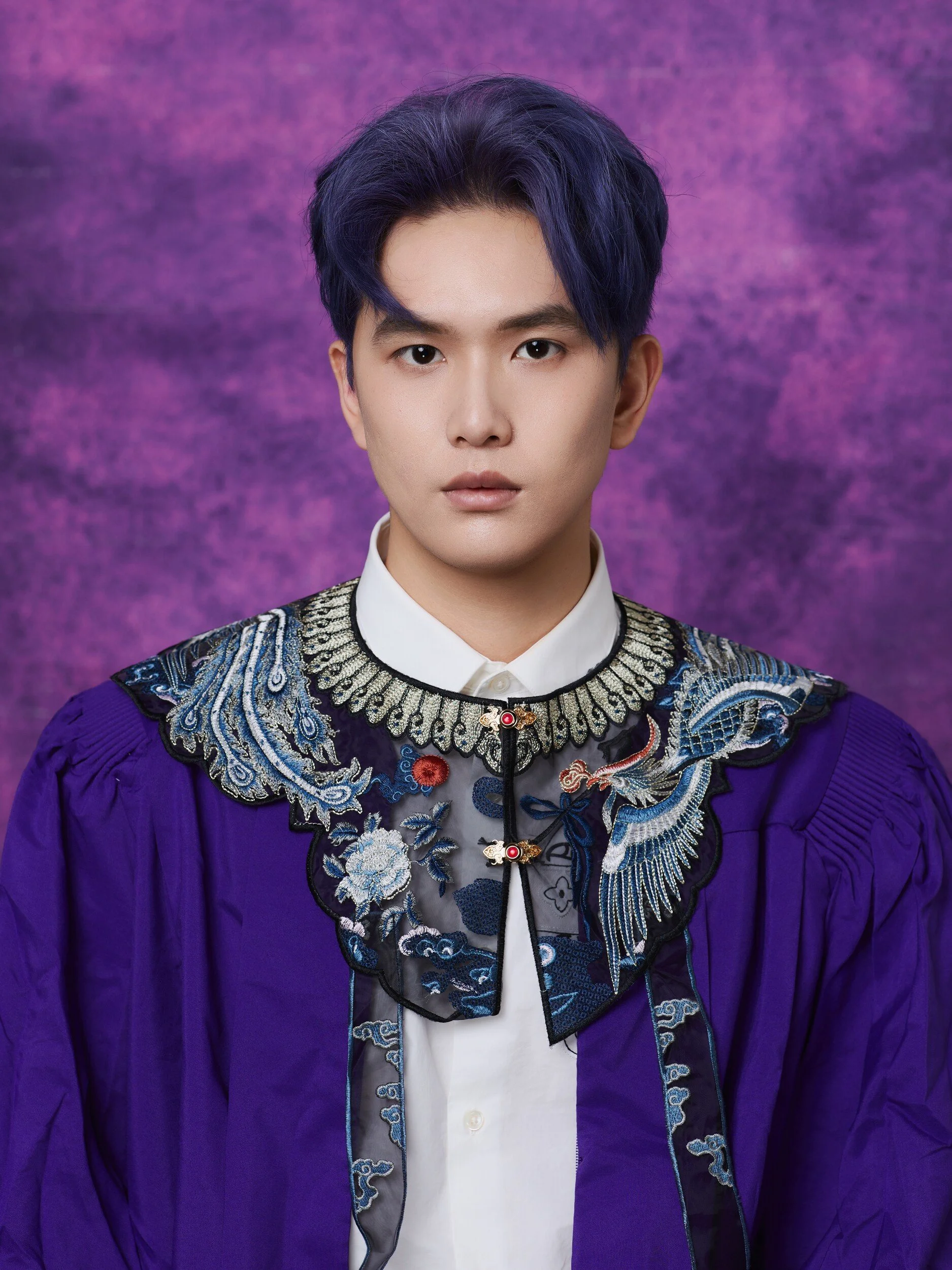 Graduation portrait of a young man with blue-toned hair in an embroidered collar under his purple gown
