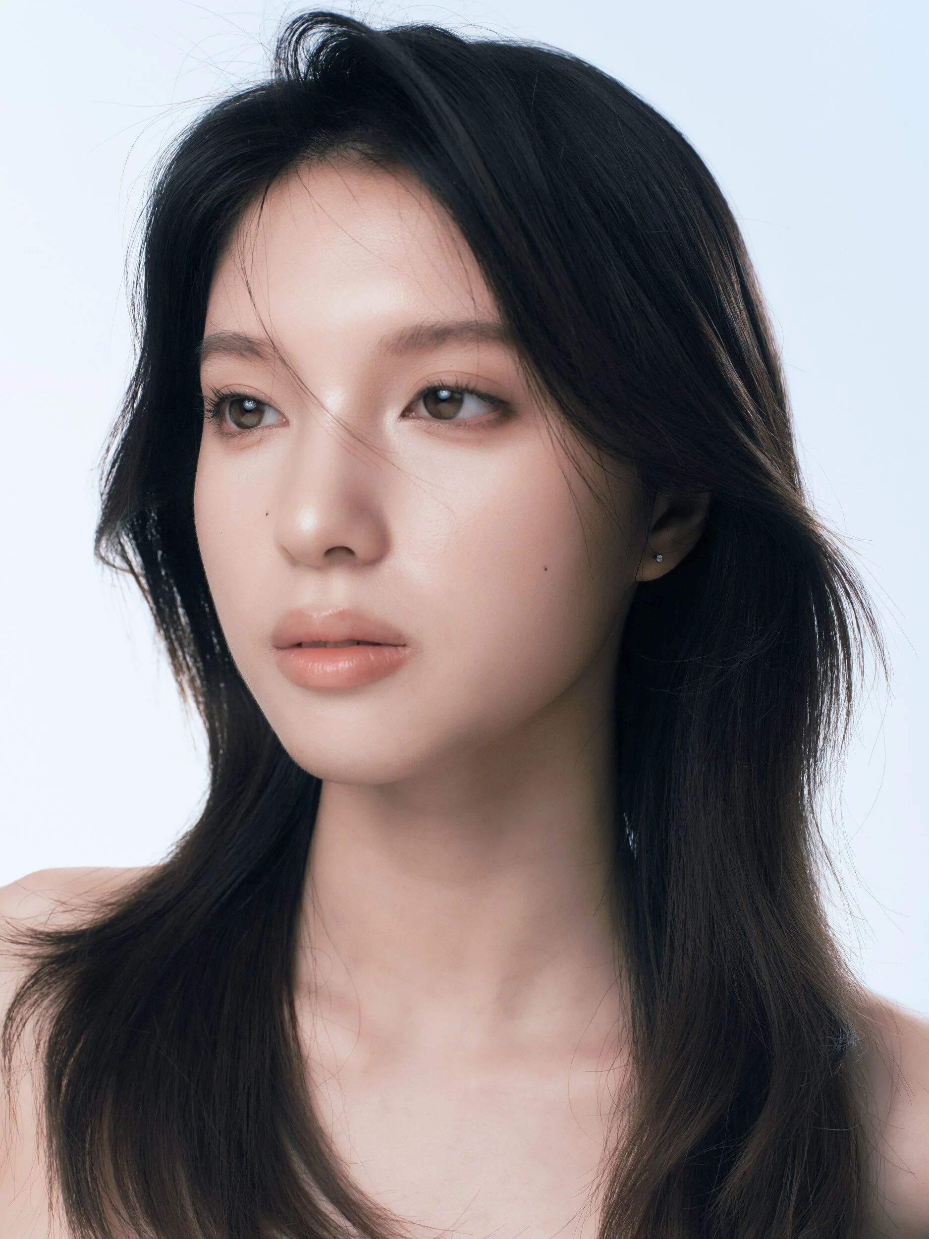 Clean beauty portrait of a woman with long layered dark hair, dewy skin and peach lips, gazing off camera on a pale blue backdrop