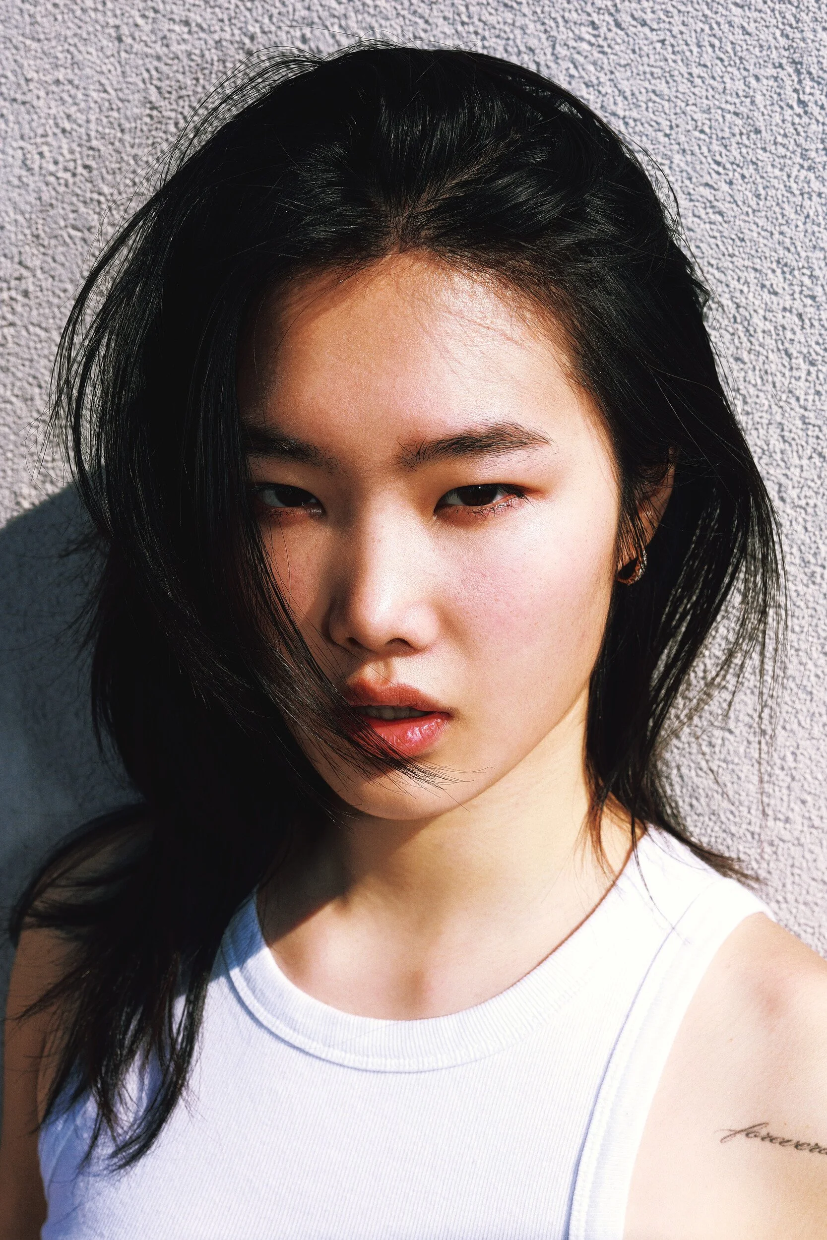 Model digitals outdoors: tight close-up of a young woman with dark hair and a white tank top, sunlit wall behind