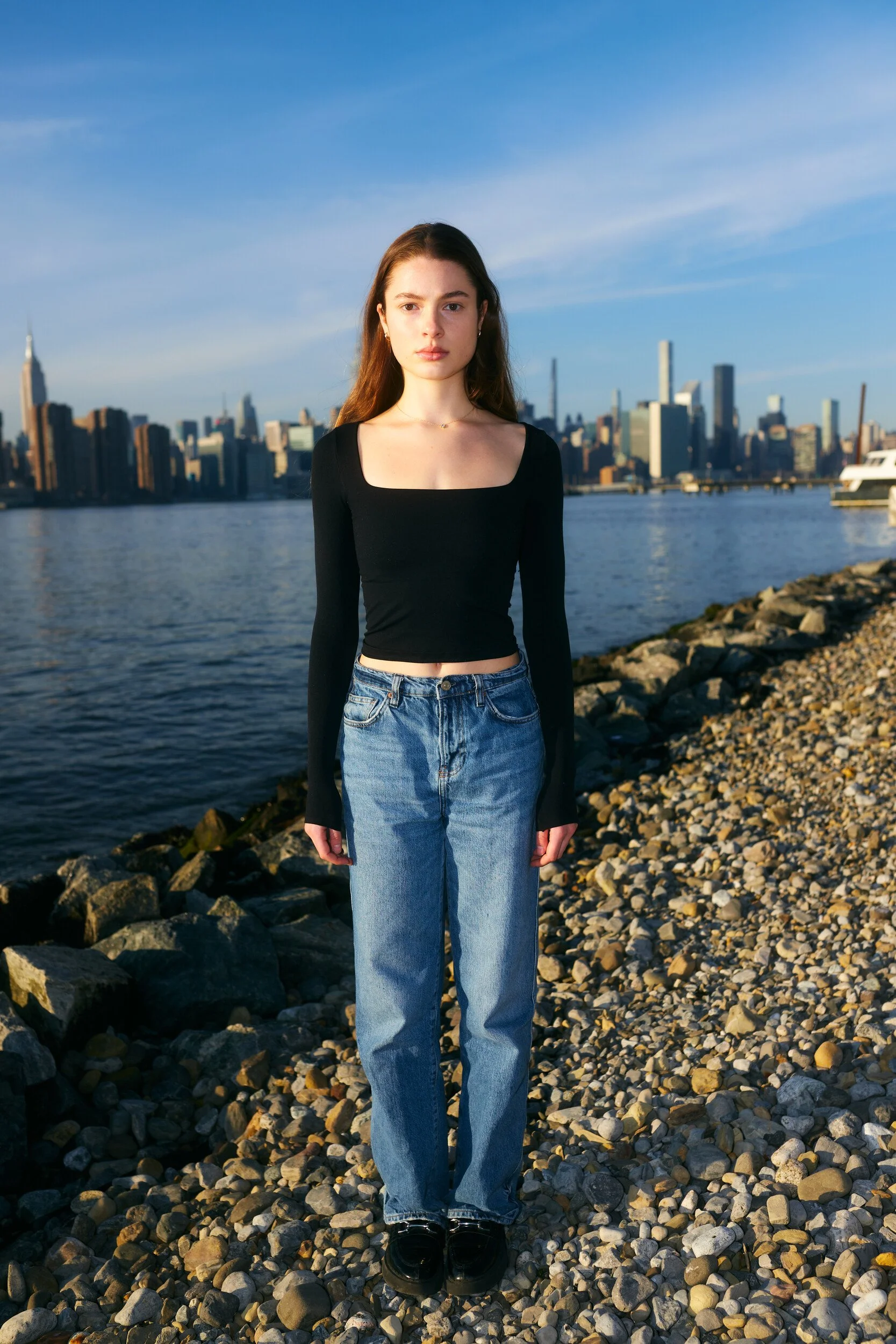 Model digitals by the river: full-length front view of a woman in a black top and jeans, Manhattan skyline behind