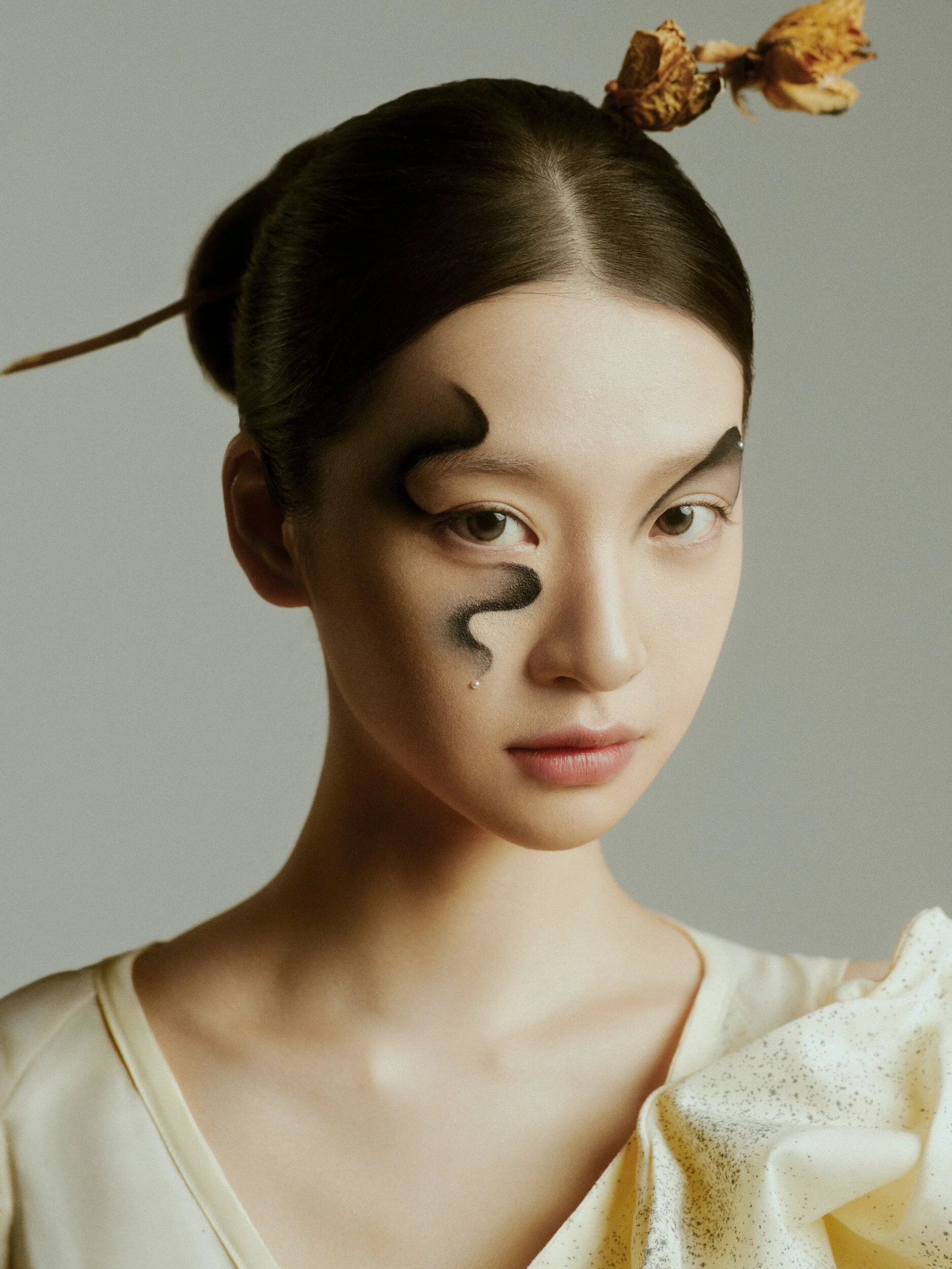 Beauty portrait with painted ink-wave face art across the eye and cheek, hair in a low bun with a dried-flower hairpin