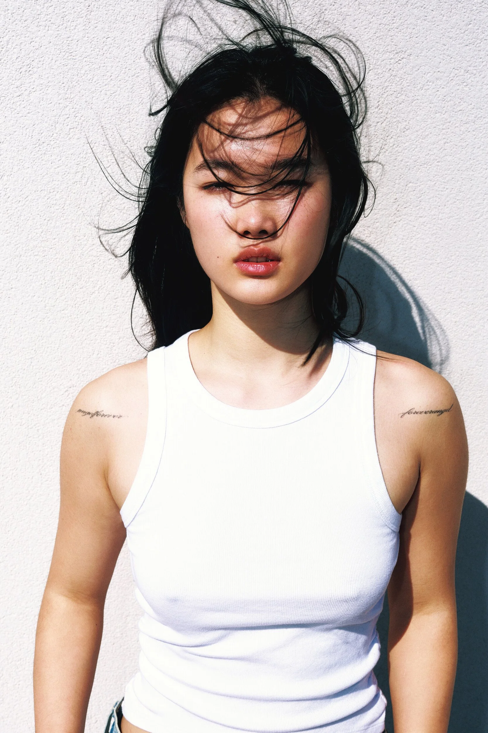 Model digitals outdoors: close-up of a young woman in a white tank top with windblown dark hair against a sunlit wall